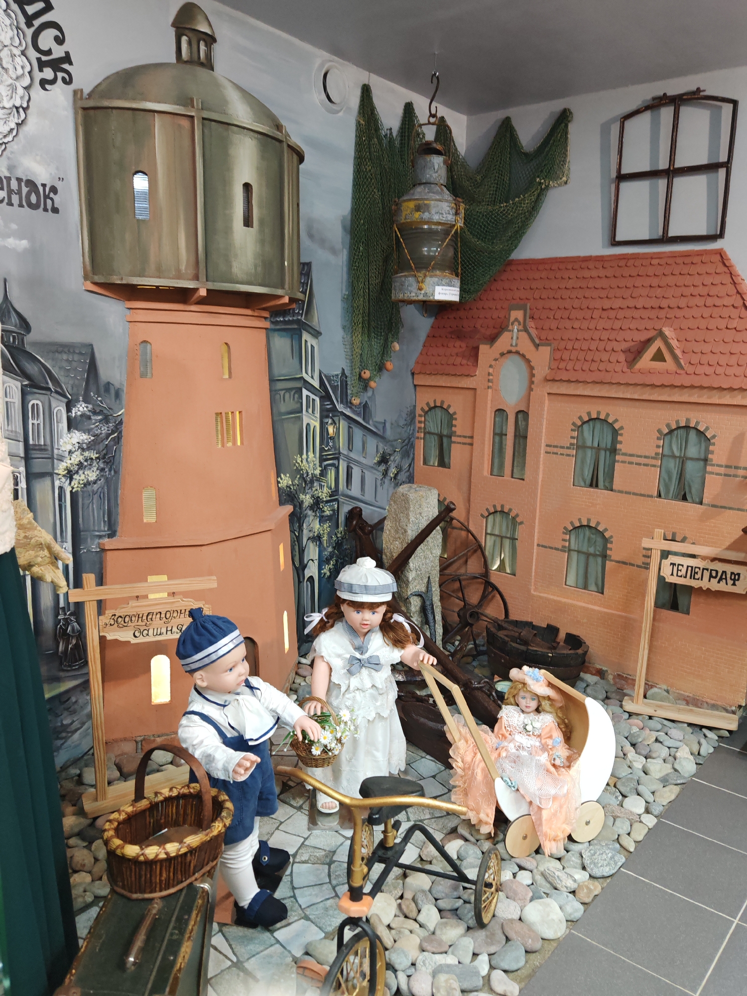 A staged scene depicts a vintage display with dolls and props set against a painted mural of a European town. In the foreground, two porcelain dolls are positioned. One doll, dressed as a sailor boy with a blue and white striped hat and overalls, stands to the left, holding a small basket with white flowers. A second doll, a girl in a white lace dress and matching hat, stands to the right, pushing a vintage doll stroller containing another baby doll dressed in a peach-colored outfit.
Behind the dolls, a large, terracotta-colored tower, resembling a lighthouse or water tower, dominates the left side of the frame. A wooden sign with Cyrillic text, possibly indicating a location, is attached to a post in front of the tower. To the right of the tower, a brick building with red tiled roofs and arched windows is depicted. A sign on this building reads "ТЕЛЕГРАФ" (Telegraph) in Cyrillic.
Other elements in the scene include a large, antique lamp hanging from the ceiling, fishing nets draped on the wall, a wooden barrel, and parts of antique machinery, including large wheels. The ground is covered with a mix of small stones and patterned tiles, suggesting an outdoor or cobblestone street setting. The overall impression is a diorama or museum exhibit designed to evoke a historical or maritime atmosphere.
