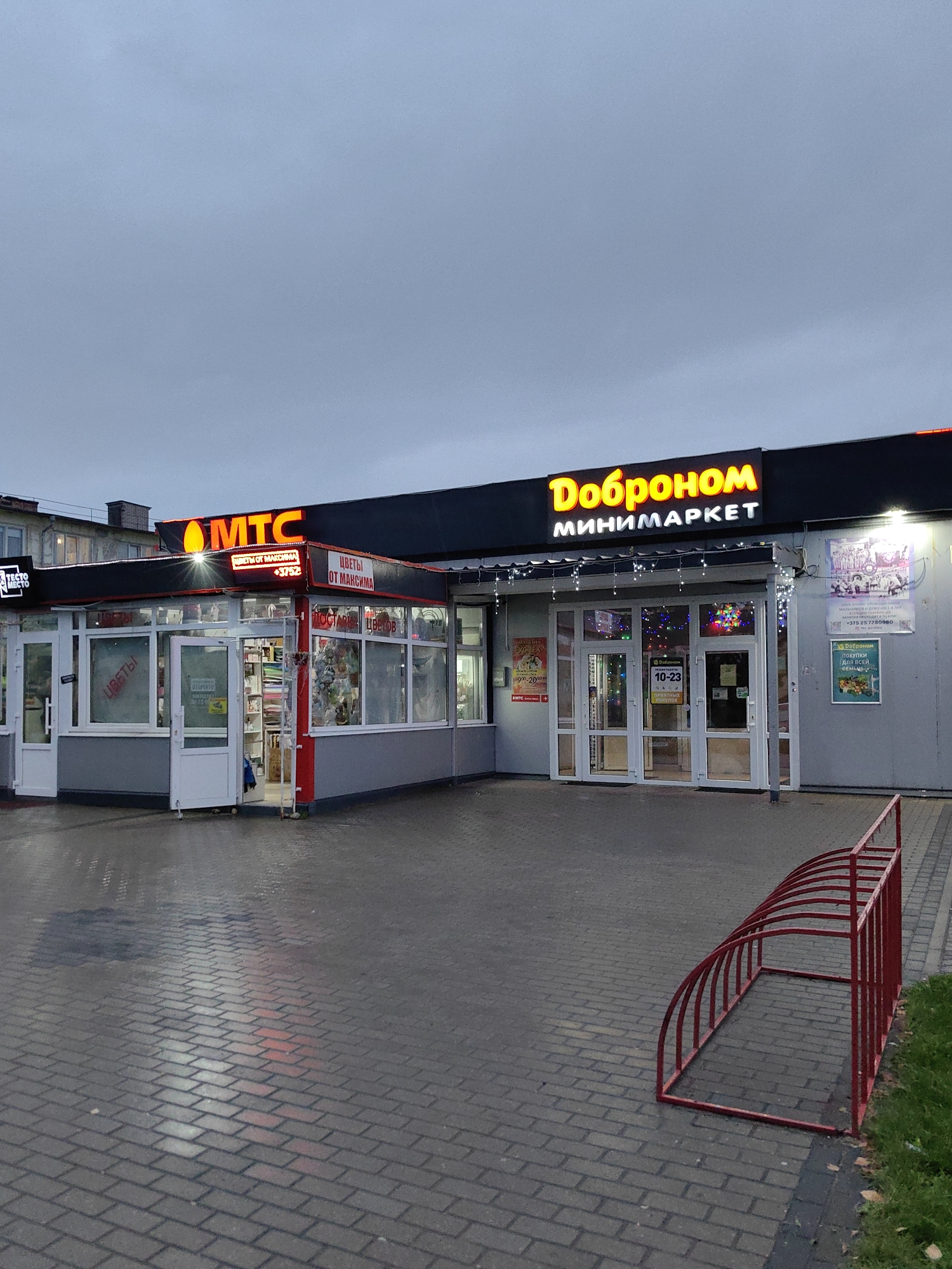 The image shows a street view of a shopping area in the city of Fanipal, Belarus. Two storefronts are visible: one is an MTS (mobile network operator) shop, indicated by a prominent "MTC" sign. This shop appears to sell flowers, as suggested by the signage "ЦВЕТЫ ОТ МАКСИМА" (Flowers from Maxima) and "ДОСТАВКА ЦВЕТОВ" (Flower Delivery). The other storefront is a minimarket named "Доброном" (Dobronom), with a large illuminated sign.
The minimarket has a modern glass facade with white framing and is decorated with string lights. Inside, shelves stocked with merchandise are visible. A poster on the wall of the minimarket advertises a "Доброном для всей семьи" (Dobronom for the whole family). Another poster, partially visible on the MTS shop, displays a phone number and possibly flower prices.
The ground in front of the shops is paved with grey interlocking bricks, which appear wet, suggesting recent rain. A red metal bike rack, shaped like a curved arch with vertical supports, is positioned on the right side of the frame. The sky is overcast and grey, indicating a cloudy day. No people are clearly visible in the scene. The overall scene depicts a commercial street in a European town, likely during the daytime or early evening due to the illuminated signs.