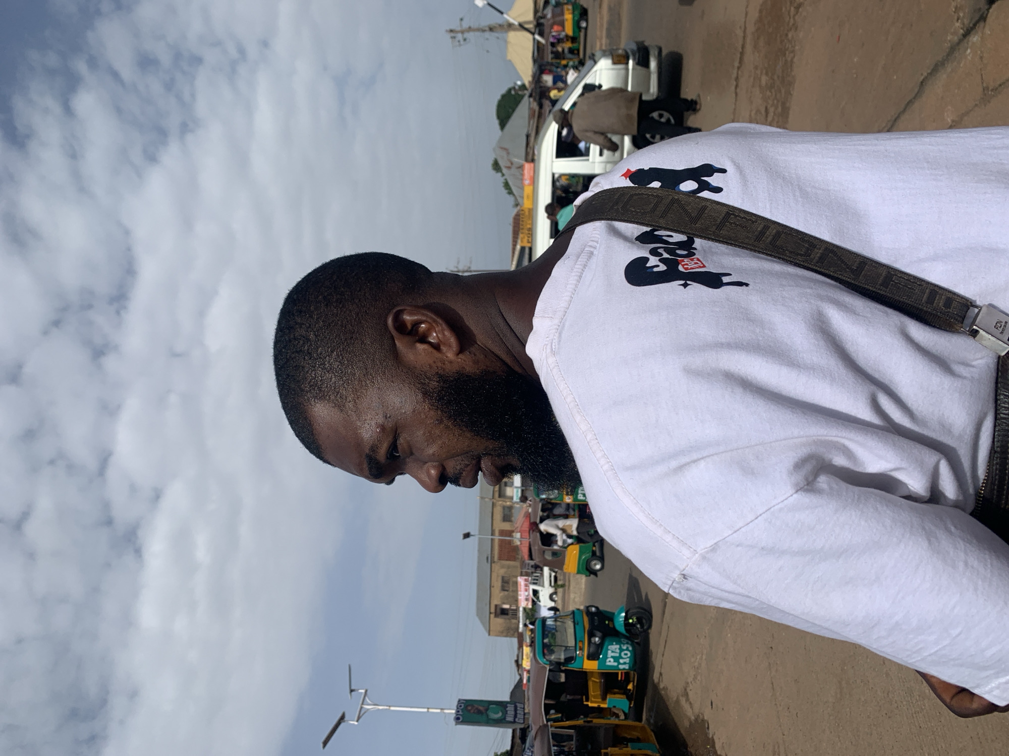 In the city of Gura Top, Nigeria, a Black man with a beard and short haircut stands facing left, looking away from the camera. He is wearing a white t-shirt with a graphic design on the back, featuring the word "Faad" in a dripping font, and a dark brown strap with a repeating logo pattern across his shoulder. The man is standing on a dusty, paved surface, likely a street, with several auto-rickshaws and a white van in the background. The sky is partly cloudy. The scene appears to be a busy urban environment with market stalls and buildings visible.