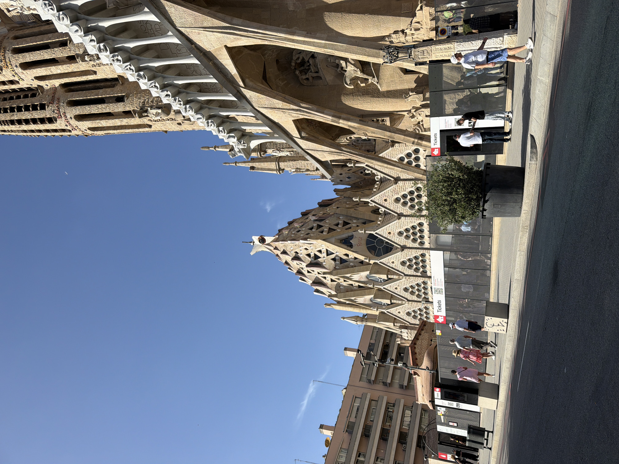 The image shows the Sagrada Familia in Barcelona, Spain. The Basilica's distinct facade, characterized by intricate stone carvings and towering spires, dominates the right side of the frame. Construction barriers with "Tickets" signs are visible in front of the building, with several people walking along the sidewalk. One person in the foreground is wearing blue shorts and a white t-shirt, walking towards the right. A large black planter with a green shrub sits on the sidewalk to the left of the ticket booth. A residential building can be seen on the left side of the image, under a clear blue sky. The street in front of the sidewalk appears to be asphalt.