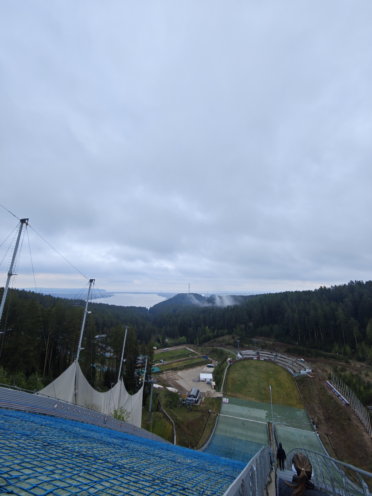The image shows a view from the top of a ski jump in Прикамский, Russia. The foreground is dominated by the blue artificial turf of the jump. Metal support structures and a white fabric screen are visible on the left. Two individuals are descending the stairs beside the jump. Below the jump, there is a grassy landing area surrounded by a small stadium seating area, along with various buildings and support structures. In the background, a dense forest extends to the horizon. A body of water, possibly a river or lake, is visible in the distance, and a transmission tower can be seen on the far shore. The sky is overcast with grey clouds.