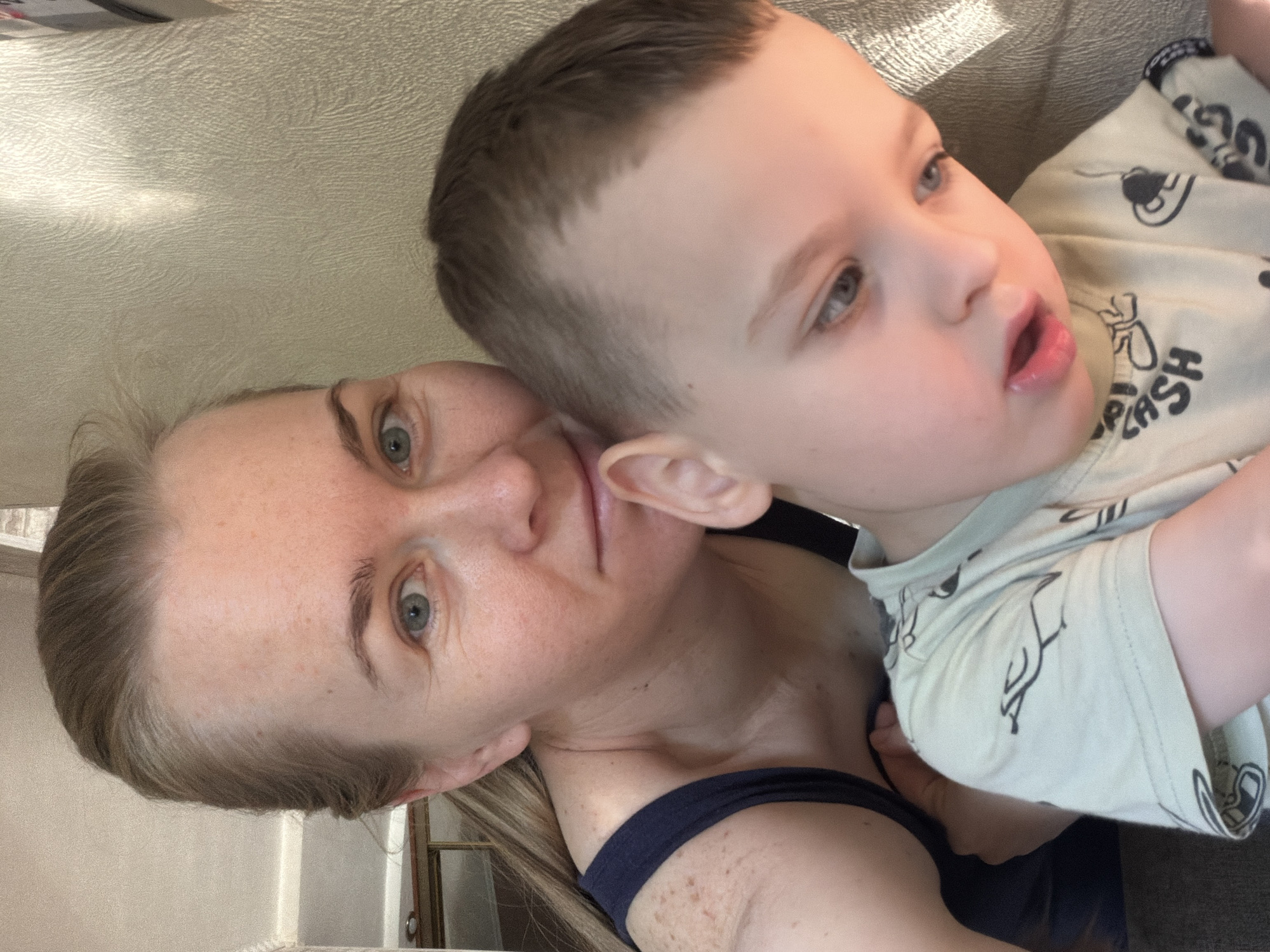 An indoor close-up selfie shows a woman and a young boy, likely in Khabarovsk, Russia. The woman, with blonde hair pulled back, has a light complexion with freckles on her shoulder and upper chest and is wearing a dark blue tank top. She is looking into the camera, while the boy, in the foreground, is looking down and to his right. The boy has light brown hair and is wearing a light-colored t-shirt with a black graphic print. They are positioned in front of a textured, light-colored wall. The lighting suggests natural light coming from the right, casting some shadows. The background is blurry but hints at an indoor setting with some shelving visible on the left.