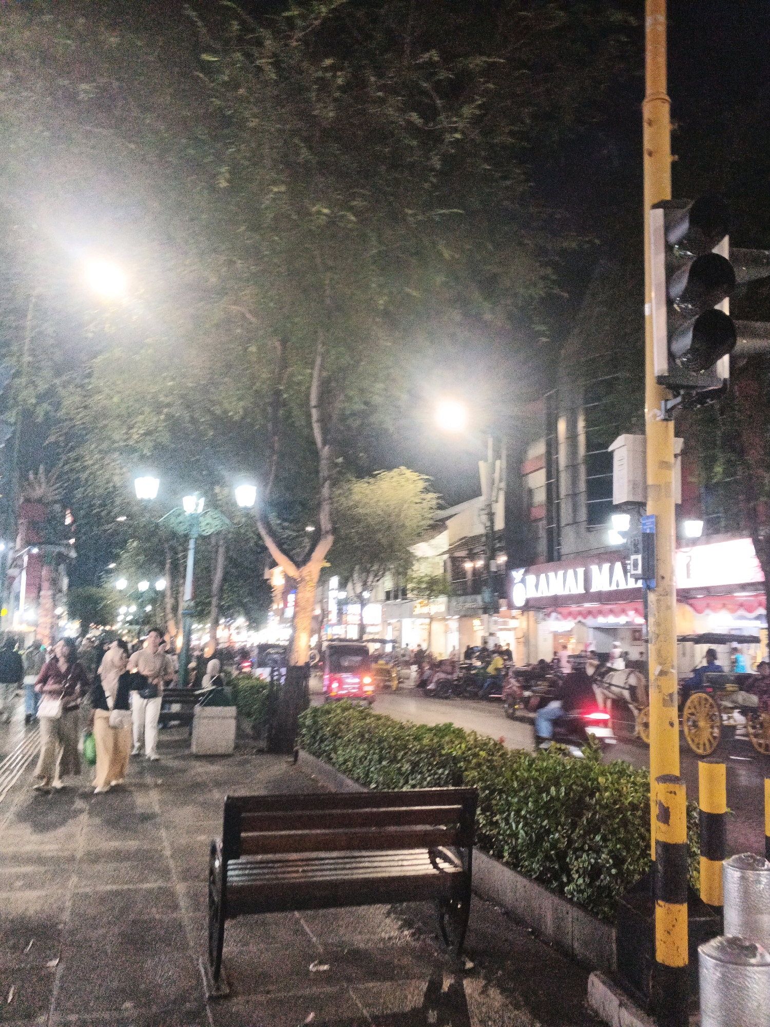 This is a nighttime street scene in Yogyakarta, Indonesia, likely on Malioboro Street. The image depicts a bustling pedestrian area with several people walking on a paved sidewalk lined with trees and streetlights. A dark wooden bench sits prominently in the foreground. Across the street, commercial buildings with illuminated signage are visible, including one with "RAMAI MALIOBORO" written in large letters. Various vehicles, including motorbikes and horse-drawn carriages, are present on the street, suggesting moderate traffic. The overall atmosphere is lively and urban, illuminated by artificial lights from streetlamps and buildings, with a dark, clear night sky.