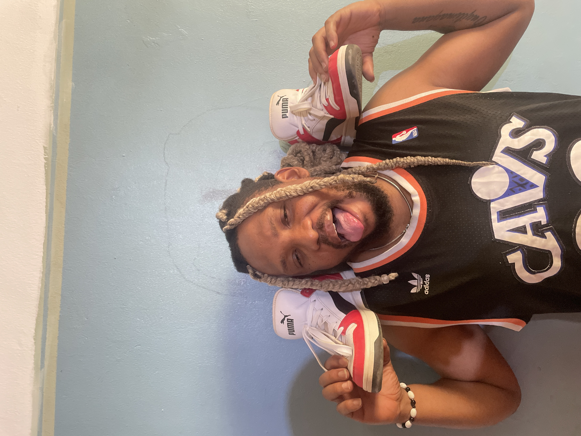 A person wearing a black Cleveland Cavaliers Adidas basketball jersey is lying on their back against a light blue wall. They are holding a pair of Puma sneakers on their head and are sticking their tongue out. The person has braided hair and is wearing a silver chain necklace and a beaded bracelet. The image provides no indication of the location being in Jos, Nigeria.