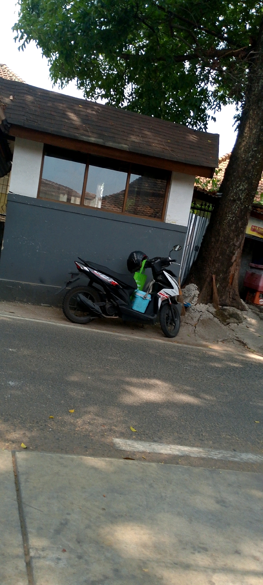 A black and white automatic scooter is parked on the side of a road in front of a building with a gray facade and a dark brown tiled roof. A black helmet rests on the seat of the scooter, and a bright green bag is behind it, with a light blue cooler box attached to the side. The building has a long, horizontal window with multiple panes, reflecting the surrounding environment. To the right of the building, a large tree with a rough bark texture is visible, and behind the scooter, there's a corrugated metal barrier. The road is asphalted, with some fallen leaves scattered on it and dappled sunlight casting shadows. The scene appears to be in a moderately urban or suburban area in Bandung City, Indonesia, given the architectural style and the presence of a scooter, which are common modes of transportation there.
