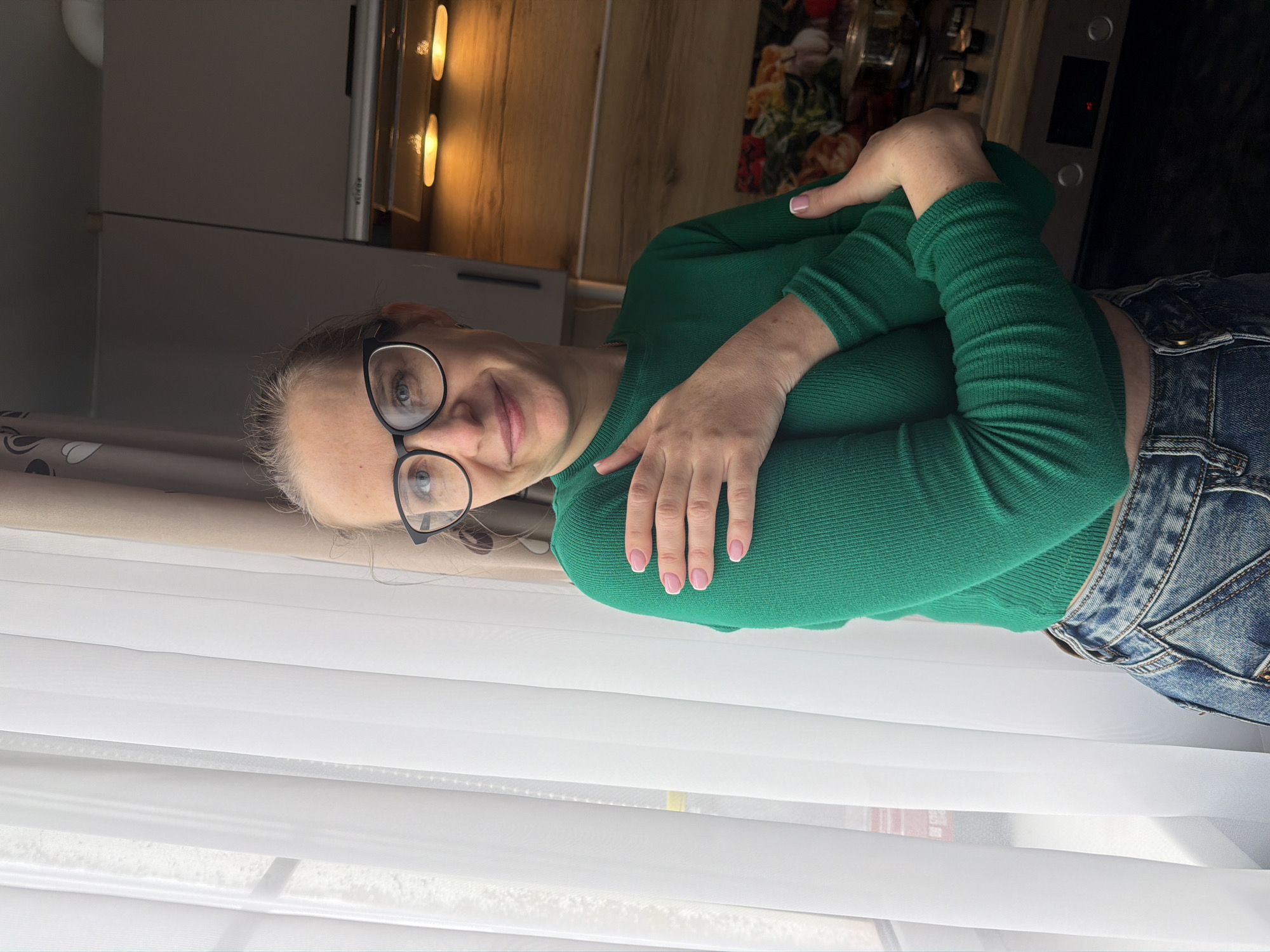 The image features a fair-skinned woman with blonde hair pulled back, wearing glasses with black frames, a green long-sleeved ribbed shirt, and blue jeans. She is posed with her arms crossed in front of her, hands near her shoulders. Her nails appear to be painted light pink.
The background consists of a kitchen setting with a white cabinet, a countertop with built-in lights, and a partial view of a stove. The lower portion of the frame shows a white sheer curtain.
There is no verifiable evidence in the image to confirm the location as Zaprudnya, Russia. Therefore, the exact location cannot be determined.