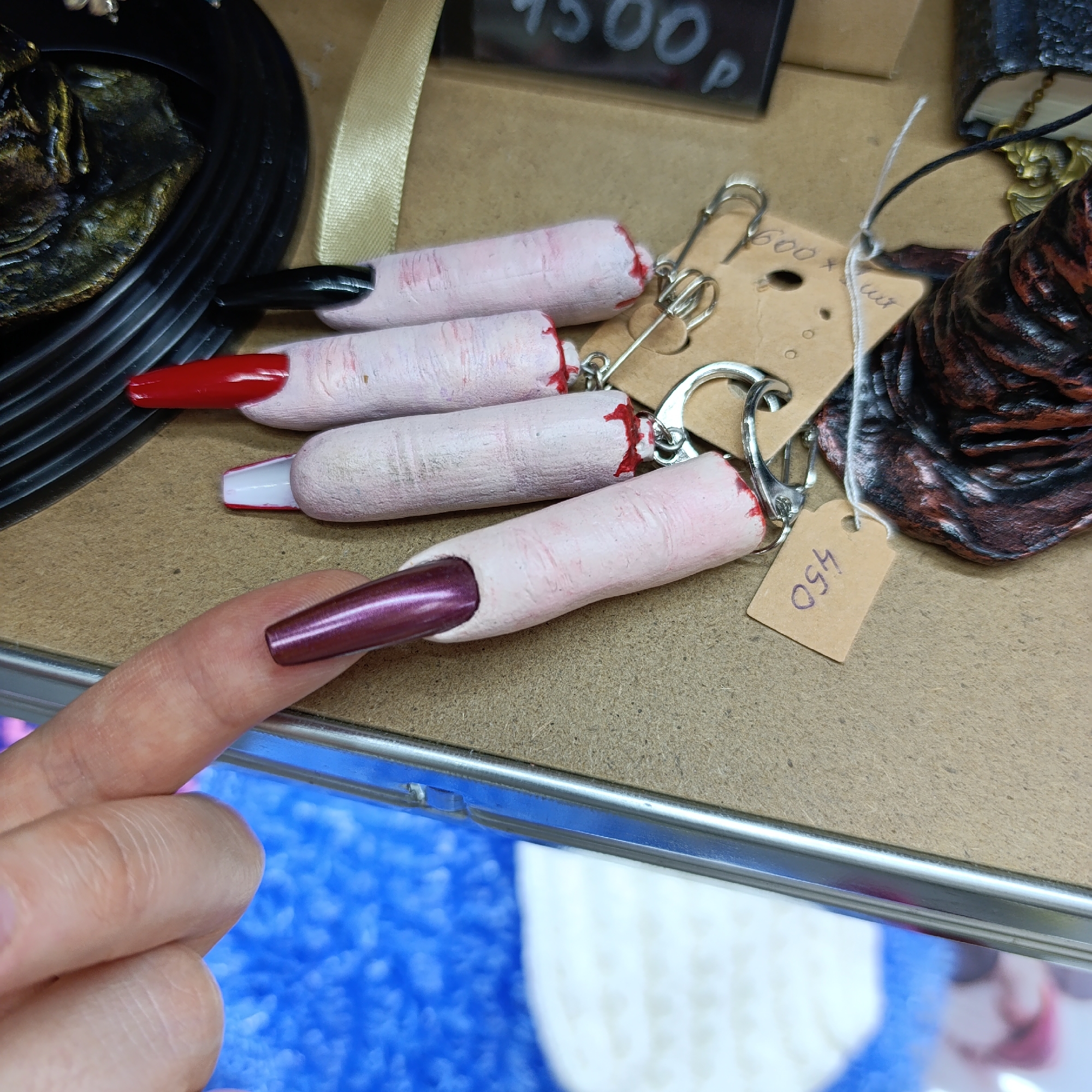 In an indoor setting, possibly a retail store or a market in Chaikovsky, Russia, a hand with a long, metallic purple nail holds a keychain shaped like a severed finger. The keychain is painted to resemble a finger with pale skin and red "blood" at the cut end. Several other similar finger keychains are on display, with nail colors including black, red, and white. They are attached to metal keyrings or clips and have price tags attached. Other merchandise is visible in the background, including a black decorative item, a ribbon, a price tag reading "4500 P," and a brown, textured object. The underlying surface is a plain beige, and the lower portion of the image shows a blurry blue and white background, suggesting more merchandise or the store's interior.