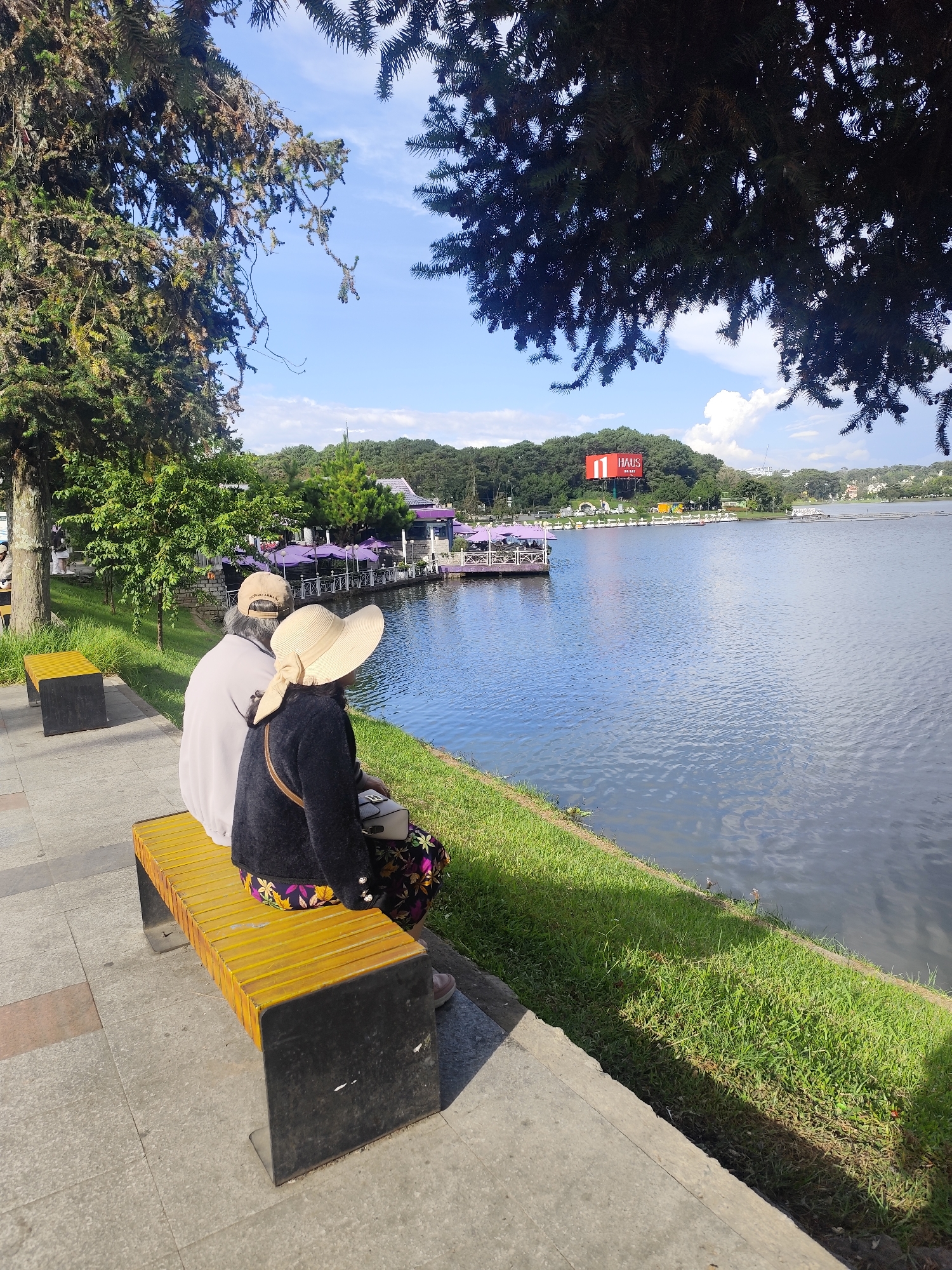Two individuals are seated on a yellow slatted bench facing a body of water in Xuan Huong Lake, Da Lat, Vietnam. The person on the left, wearing a light-colored long-sleeved shirt and a beige cap, is mostly obscured from view. The person on the right wears a black cardigan and a patterned skirt, with a wide-brimmed straw hat. A purse is placed on the bench between them. A paved walkway runs alongside the lake, bordered by a strip of green grass. In the background, across the lake, is a forested hillside with buildings and a large red billboard displaying "11 HAUS". Several purple umbrellas are visible near a waterfront structure. Trees with dense foliage frame the top of the image. The sky is blue with scattered white clouds, and the sun casts shadows on the ground.