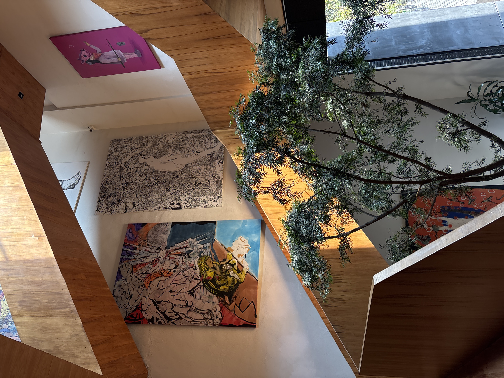 The photograph is taken from a low angle, upward, showcasing an interior space adorned with artwork and architectural elements. The primary focus is on a stairwell constructed with wooden beams that create angular lines across the frame.
Several paintings are mounted on the walls. One is a pink canvas with a figure lying down. Adjacent to it is a large, intricate black and white drawing that fills a significant portion of the wall space. Below that is a vibrant piece with a mix of colors and a detailed depiction of figures.
A tree-like plant or decoration is present in the right side of the frame. The scene is well-lit with natural light, giving the wooden structures a warm tone. The architectural design, along with the artwork, creates a sense of contemporary style. The location of the image is in Mexico City, Mexico.