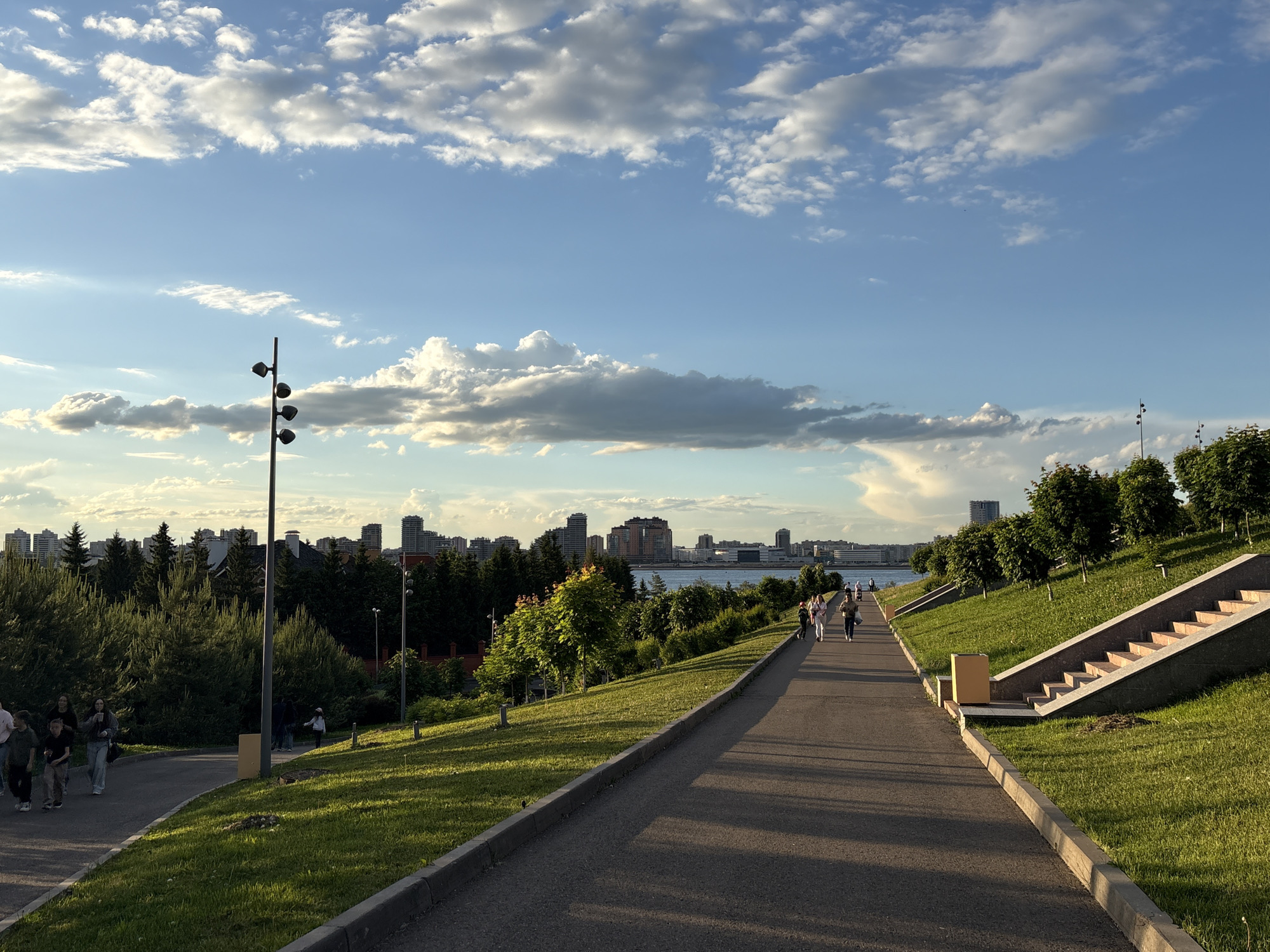 The image depicts a scenic outdoor promenade or park walkway on a partly cloudy day, likely during late afternoon or early evening. A wide, paved path runs through the center, curving slightly left in the foreground and extending into the distance, with elongated shadows stretching across it from the right. Lush green grass covers the gentle slopes flanking the path, adorned with various trees and shrubs.
Numerous individuals are visible enjoying a stroll. On a smaller path branching to the left, a group of approximately five to six people are walking. Further along the main path to the right, several other individuals or small groups are seen walking away from the viewer.
Prominently featured on the left is a tall, dark grey lamp post equipped with four spherical light fixtures. Shorter, similar light fixtures are scattered along the grassy edges of the path. To the far right, a set of concrete steps with light-colored treads ascends a steep grassy bank, next to which sits a light brown trash receptacle.
In the mid-ground, beyond the green foliage, a body of water—likely a river or large lake—is visible. A city skyline composed of numerous buildings and high-rises stretches across the distant horizon, bordering the water. The sky above is a clear blue, dotted with scattered white and grey cumulus clouds, some illuminated by the low sun. The overall scene suggests a peaceful urban green space. No visible text is present in the image.