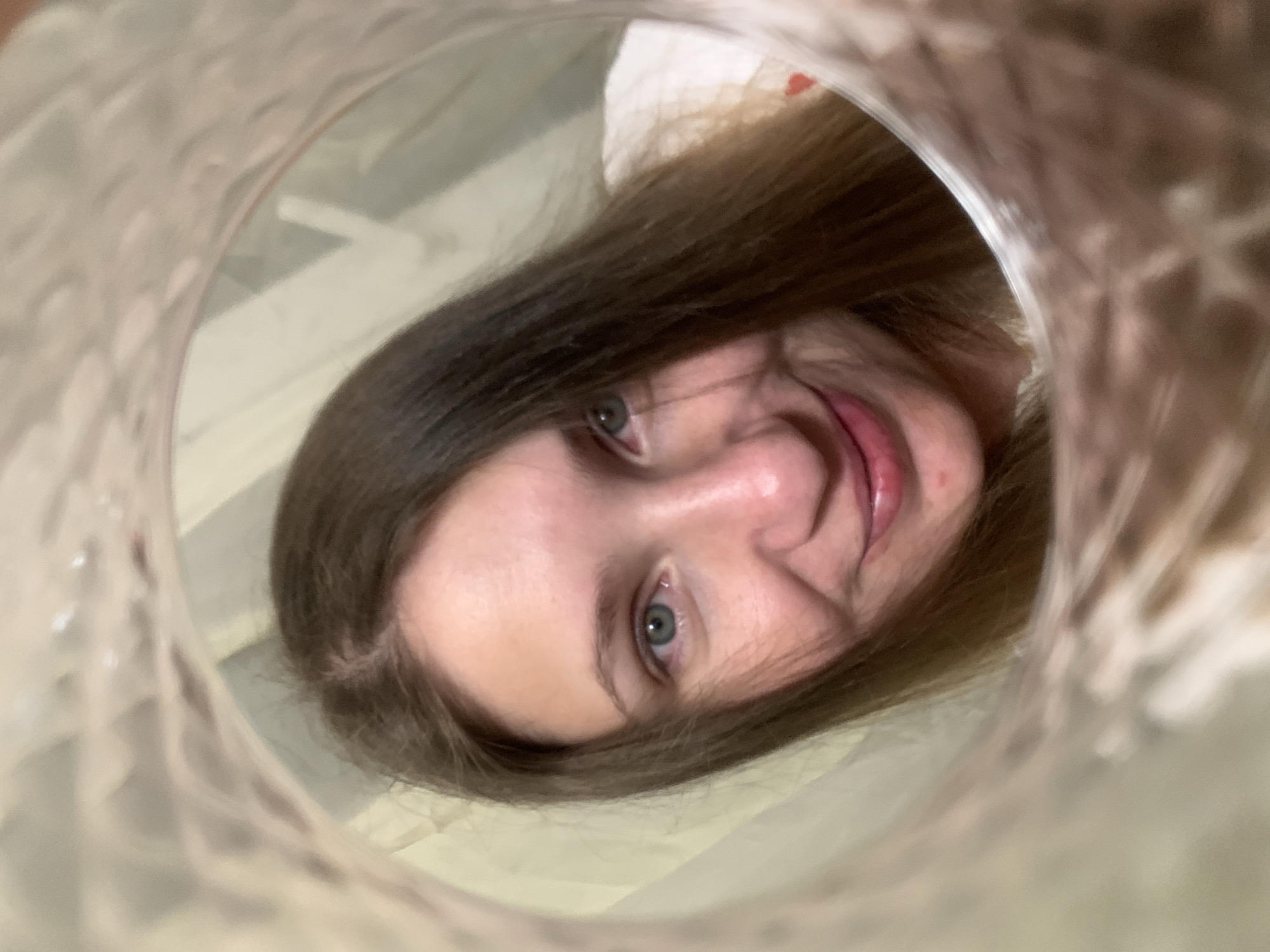 A woman's face is seen looking into a translucent plastic container, angled from above. The container creates a circular frame around her face. She has light skin, blue eyes, and brown hair. Her expression is slightly smiling. The background appears to be a blurred room interior with hints of white or light-colored surfaces. The photo appears to have been taken indoors. No location information is discernible.