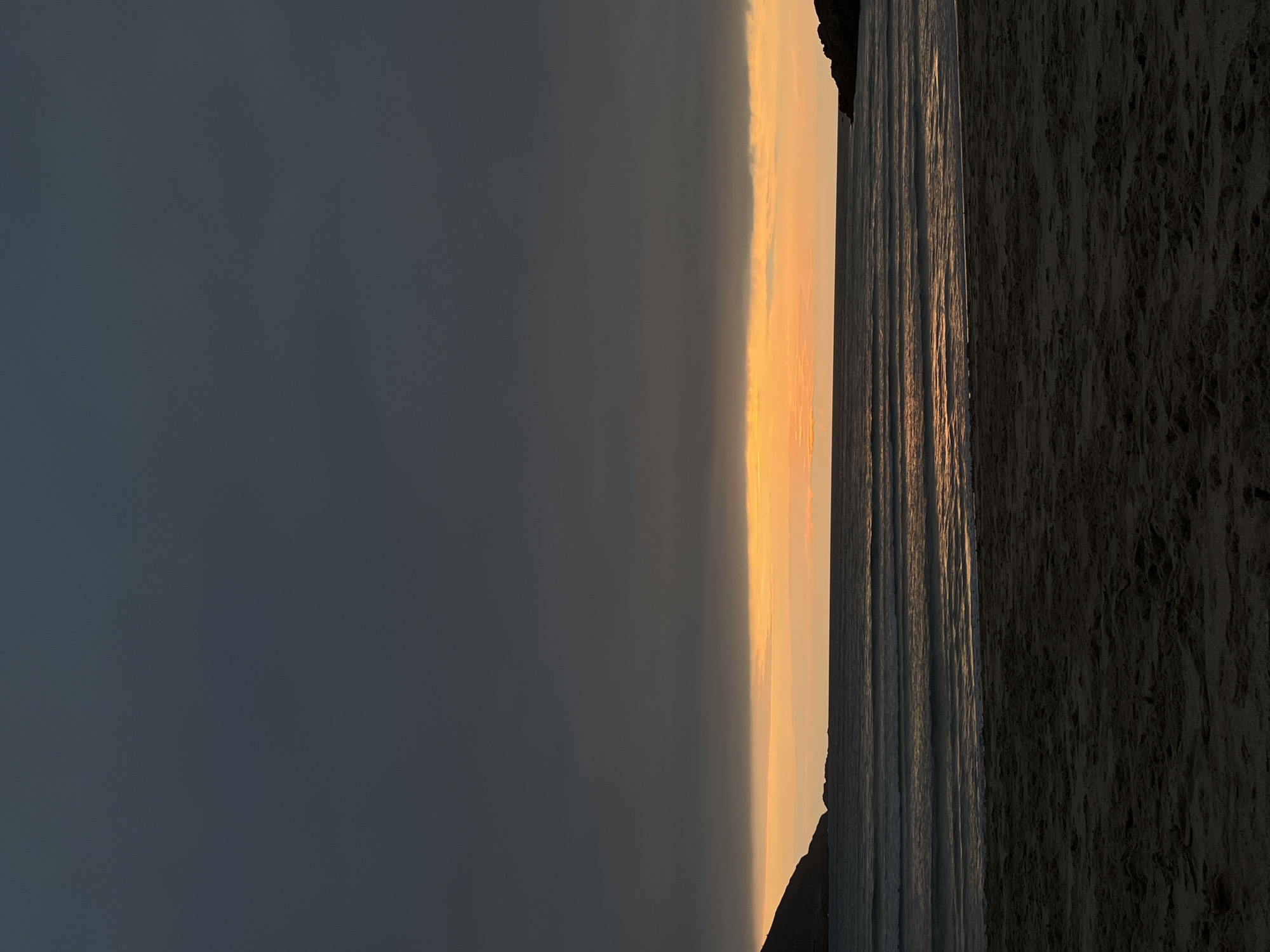 The image depicts a sunset at a beach in A Coruña, Spain. The sky on the left is dark, transitioning to a bright yellow-orange hue towards the horizon. The sea reflects the colors of the sunset with vertical lines of light. On the right, there is a dark sandy beach. A rocky outcrop is visible at the edges. The image is rotated 90 degrees.