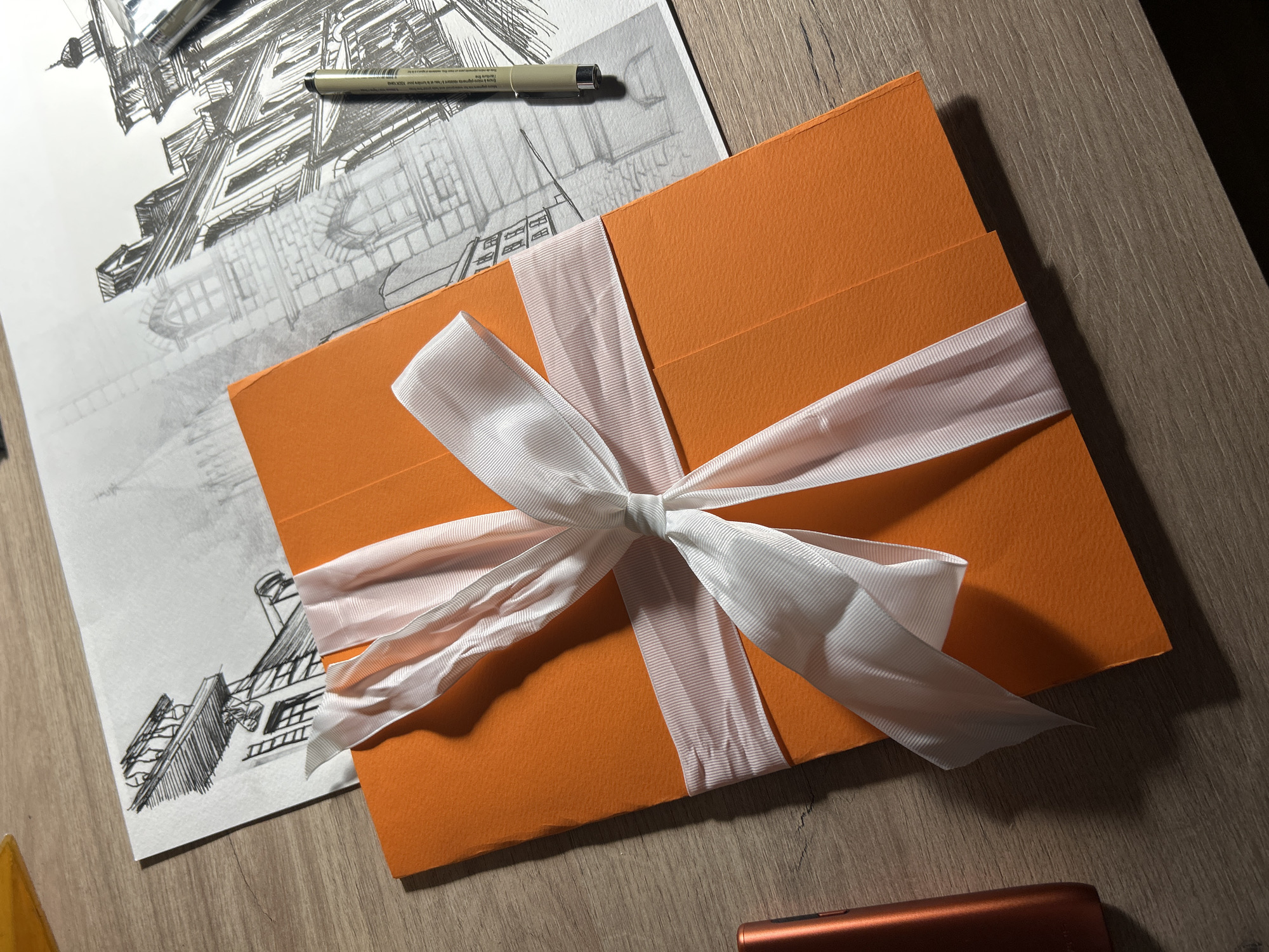 An overhead shot captures a still life arrangement on a wooden surface, likely a desk or table. The primary subject is an orange, rectangular box or folder, tied with a prominent white satin ribbon fashioned into a bow at its center. The surface of the box appears textured.
To the left and partially underneath the orange box, a detailed architectural sketch is spread on white paper. The drawing, executed in black ink, depicts an intricate building structure with multiple levels, windows, and rooflines, showcasing precise line work and cross-hatching for shading. A black fine-tipped pen, identifiable by the visible text "Pigma Micron" on its barrel, rests on the upper part of the drawing.
The wooden surface exhibits a distinct grain pattern, and ambient lighting, possibly artificial, casts soft shadows around the objects, particularly underneath the orange box and the ribbon, and to the right of the pen. A faint, reddish-brown object, possibly an electronic device, is partially visible at the bottom right corner of the frame.
The scene suggests a creative workspace or the presentation of an artistic piece, possibly a gift or portfolio. No people are visible. The time of day is not explicitly indicated but the artificial lighting implies an indoor setting, potentially evening or night. The image does not contain specific visual cues to confirm the suggested location of Sofia, Bulgaria.