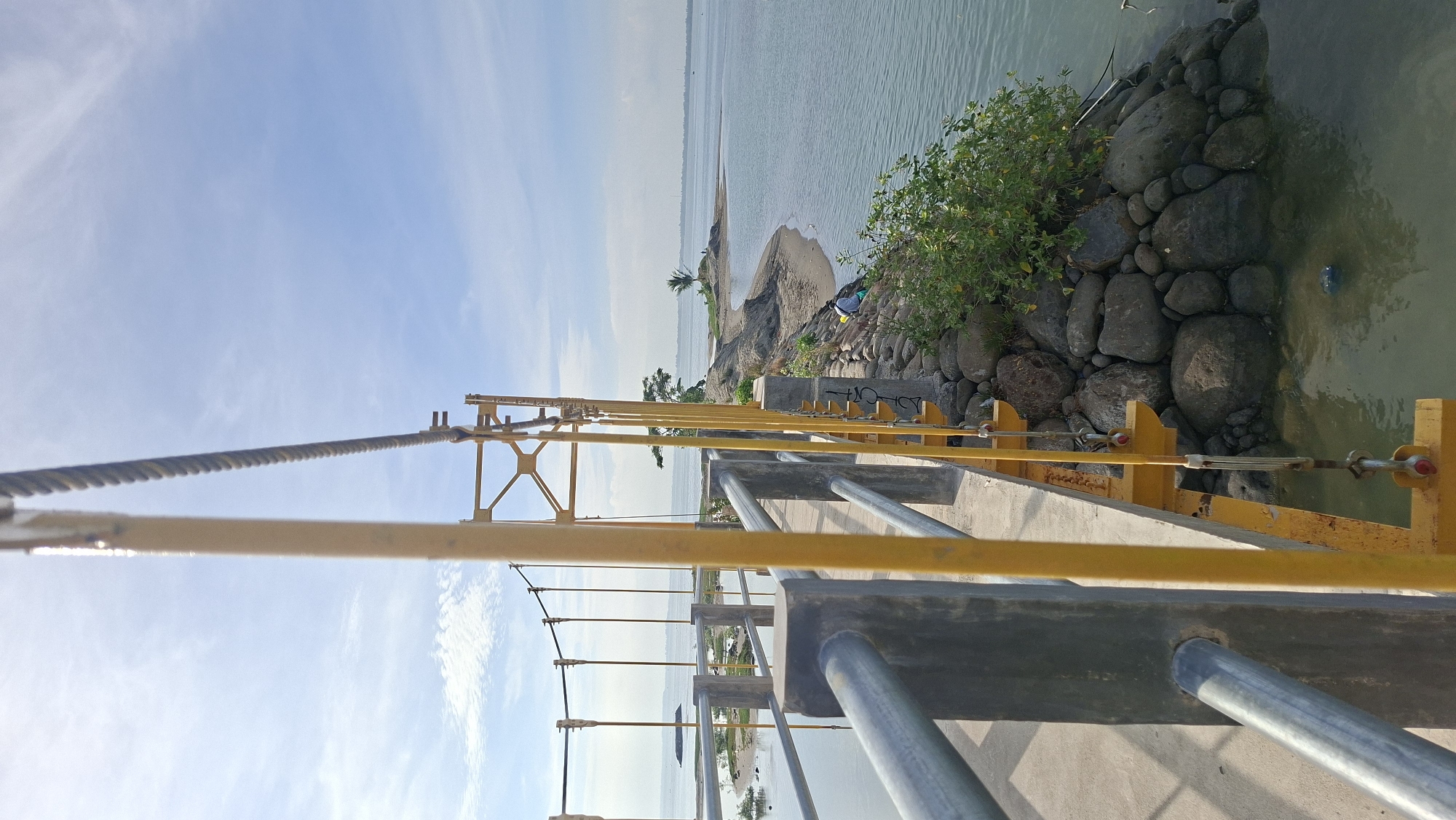 The image depicts a suspension bridge over a body of water, likely a river or a bay, in Lhokseumawe, Indonesia. The bridge has yellow support beams and silver railings. The water is calm and a light blue-green color. On the right side of the bridge, there is a rocky embankment with some green vegetation growing on it. A person wearing a light-colored shirt and dark pants is sitting on the rocks, facing the water. In the distance, a sandy peninsula with a few trees extends into the water. The sky is blue with scattered white clouds. The overall scene suggests a peaceful, natural environment within the city.