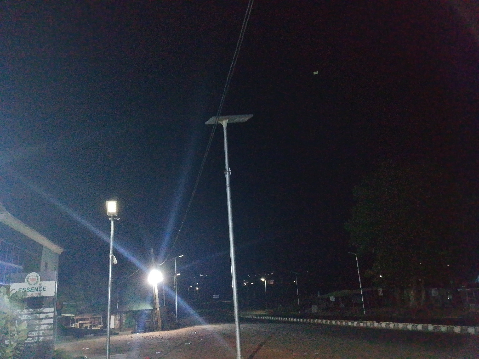 The image shows a street scene at night in Adankolo, Nigeria. Streetlights illuminate the road and surrounding area. There are buildings on the left side of the image and vegetation on the right. Power lines are visible above the road.