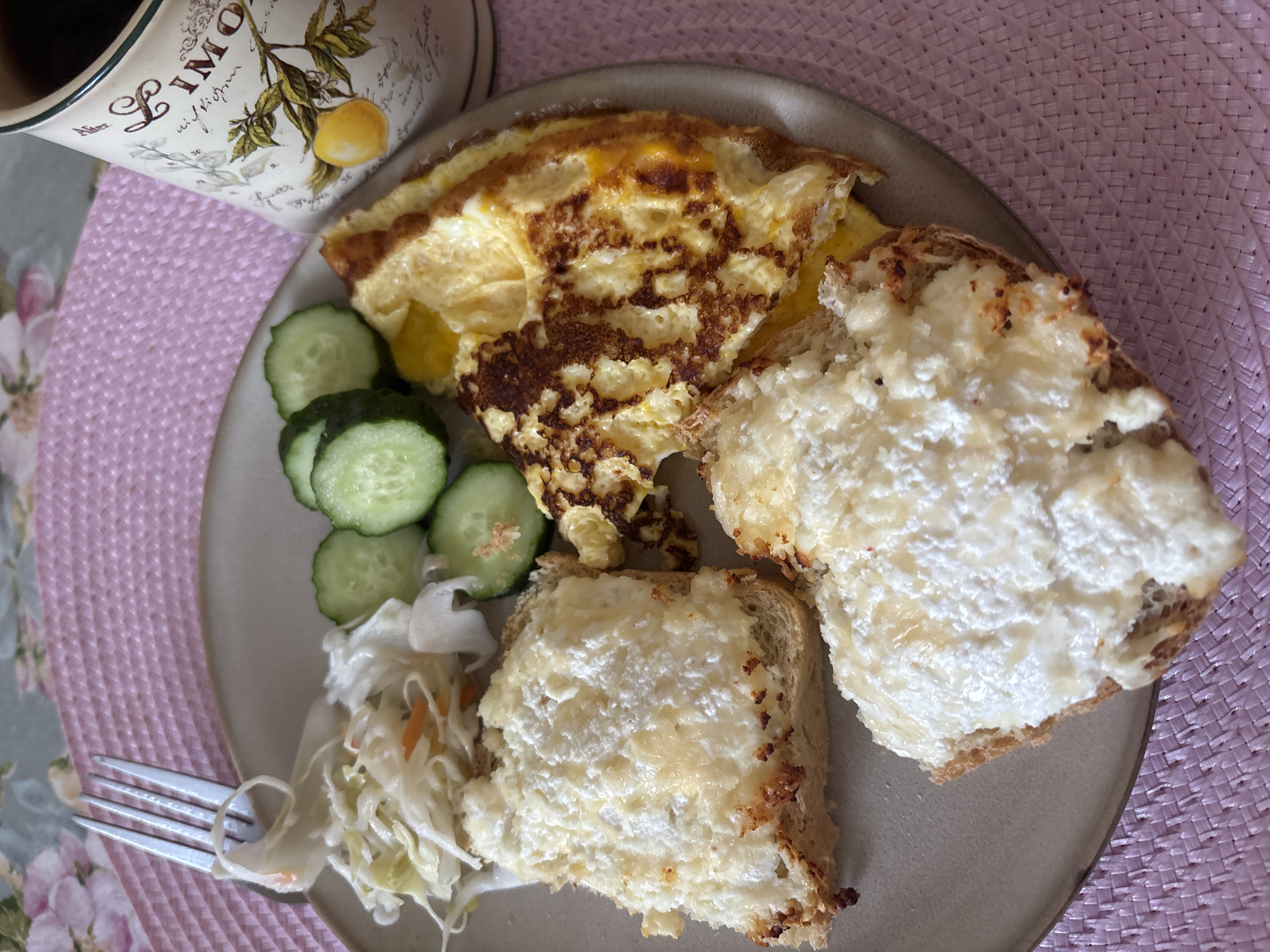 An overhead shot shows a breakfast plate on a pink woven placemat. The plate contains two slices of toast topped with a white spread, a folded omelet, cucumber slices, and a small portion of coleslaw with a fork. A mug with a lemon design is partially visible to the right of the plate. The background is a floral tablecloth. There is no indication of Naberezhnye Chelny, Russia.