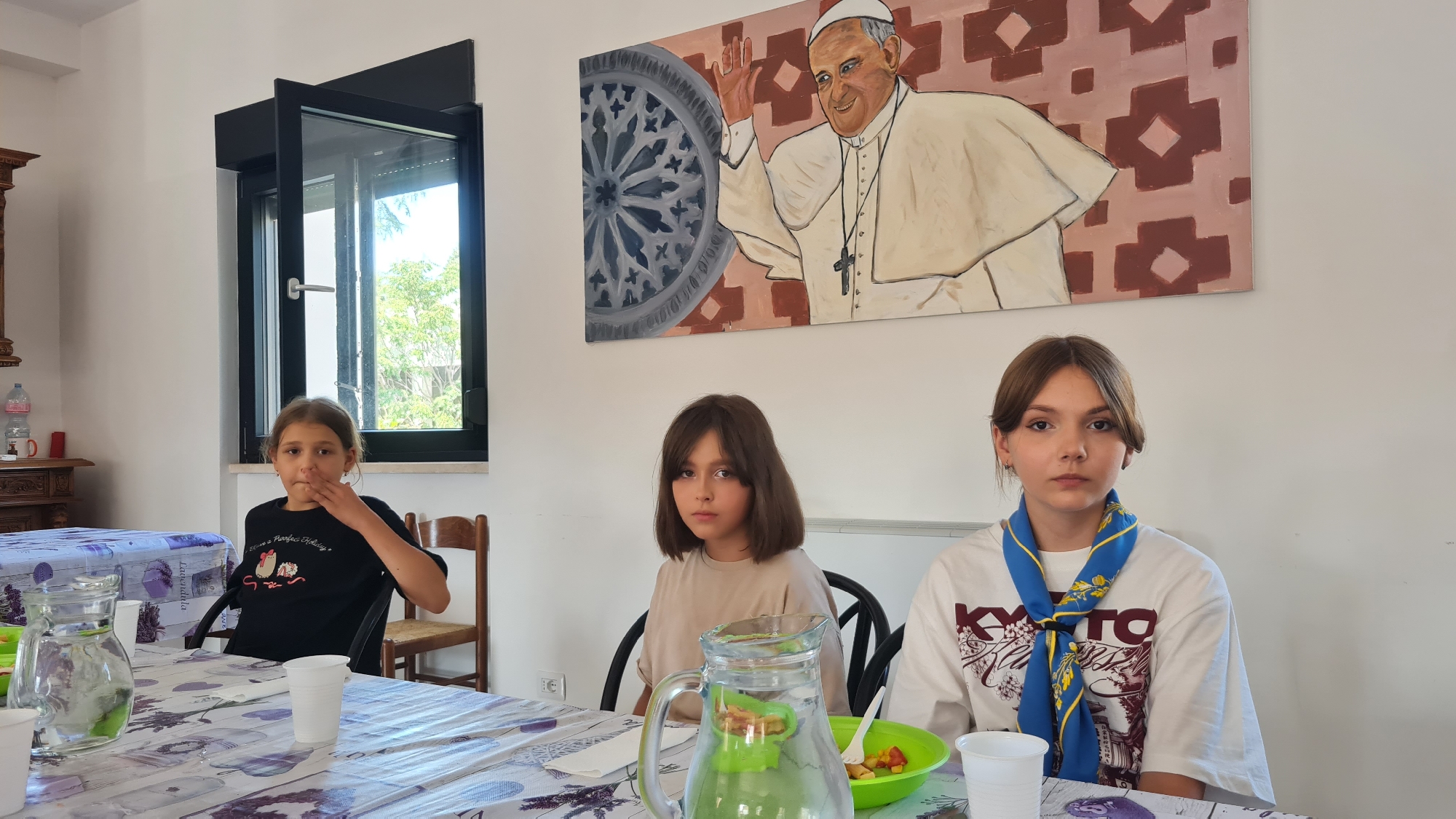 Three young girls are seated at a table in what appears to be a dining room or common area. The girl on the left, wearing a black t-shirt, has her hand near her mouth. The middle girl, in a light brown t-shirt, has a neutral expression. The girl on the right, wearing a white t-shirt and a blue and yellow scarf, looks directly at the camera. The table is covered with a patterned tablecloth and set with pitchers, cups, and a bowl of fruit salad. In the background, a large painting of Pope Francis hangs on a white wall, and a window shows trees and foliage outside. A wooden chair is visible to the left of the middle girl. The room appears to be in L'Aquila, Italy.