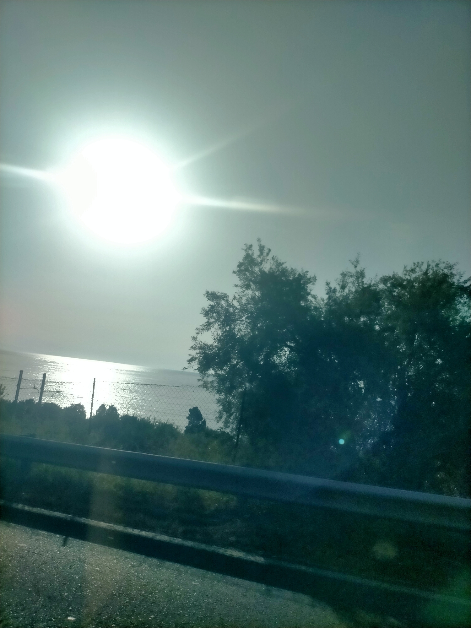 The image depicts a bright, hazy sun shining over a body of water. In the foreground, there is a road surface and a metal barrier. Behind the barrier, a fence can be seen, with some vegetation and trees beyond that. The overall scene is a landscape view, likely taken outdoors.