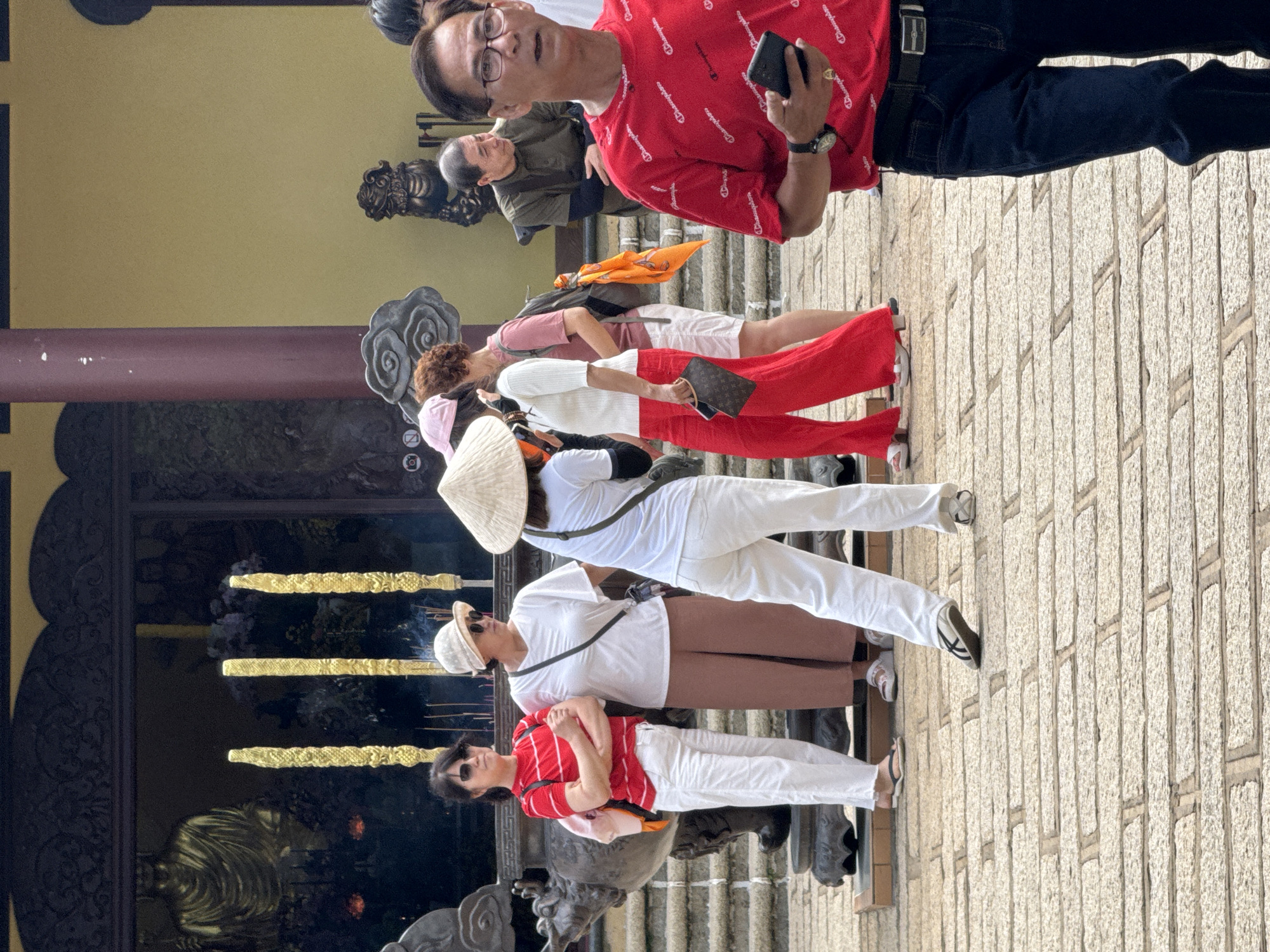 A group of people are gathered in front of a temple in Da Lat, Vietnam. The temple has a traditional architectural style with ornate carvings and golden statues. In the foreground, a man in a red t-shirt and jeans is holding a smartphone. He is wearing glasses and a watch. To his left, a woman in white pants and a red and white striped shirt is standing with her arms crossed. She is wearing sunglasses. Next to her, a woman in brown pants and a white shirt is wearing a hat and sunglasses. Behind them, several other people are visible, including a woman wearing a conical hat and a white shirt. The ground is paved with stone. Incense sticks are burning in front of the temple.