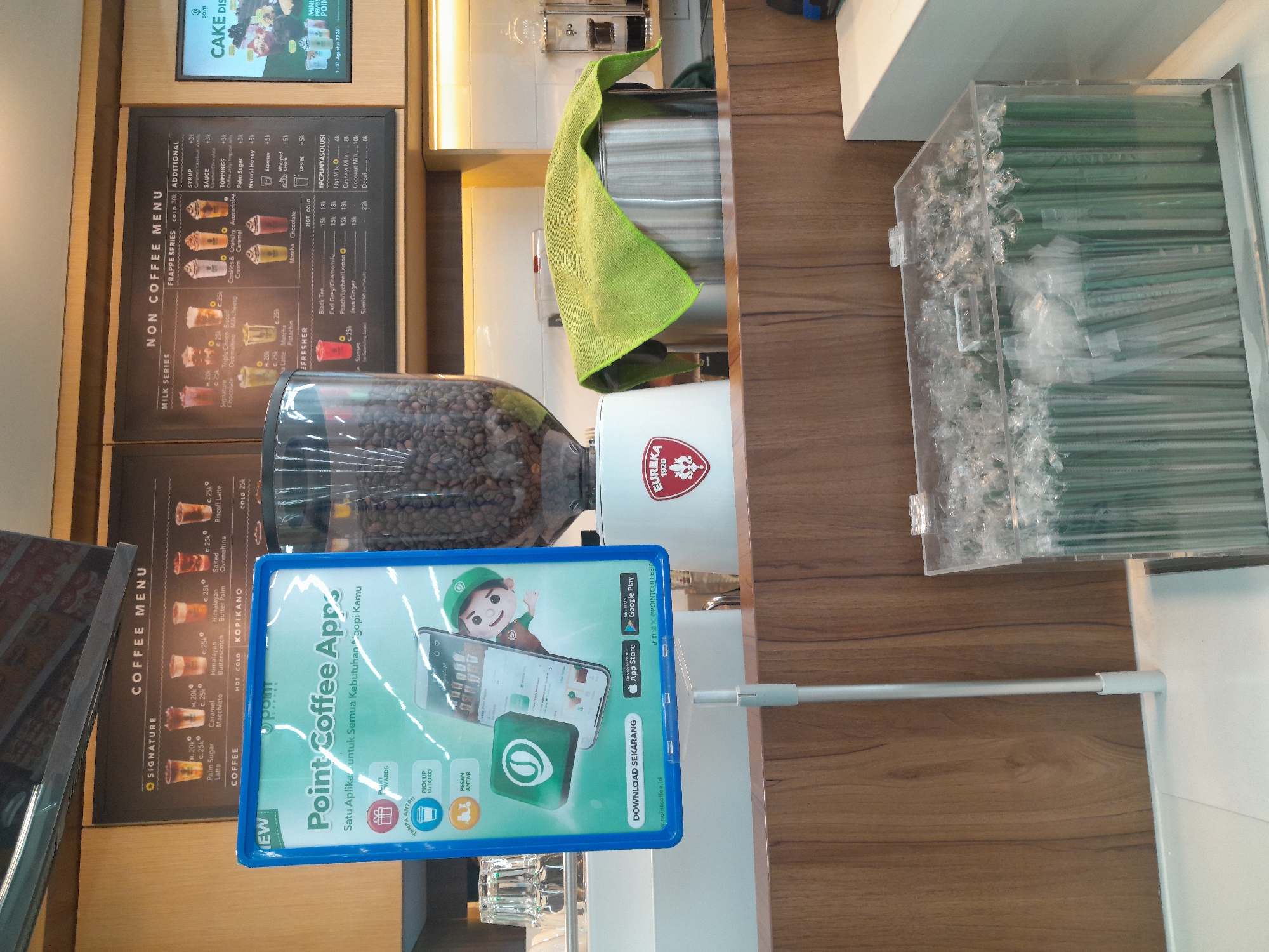 The image shows a Point Coffee shop, likely in Desa Ponokawan, Indonesia, with a focus on promotional materials and coffee-making equipment. In the foreground, a blue-framed sign advertises the "Point Coffee App," detailing its features like "Pick Up Di Toko" (Pick Up at Store) and "Pesan Antar" (Delivery Order), and encouraging downloads from the App Store and Google Play. The sign also displays the Point Coffee logo and social media handles.
Behind the sign, a large glass container filled with coffee beans sits atop a coffee grinder. To the right, a stainless steel container is covered with a green microfiber cloth, and further to the right, a digital menu board displays "CAKE DISK" and other promotional items with dates. The background features a wooden wall and shelving units.
The overall environment suggests a typical coffee shop setting, emphasizing digital ordering and customer convenience. The presence of a coffee grinder and beans highlights the core product offering.