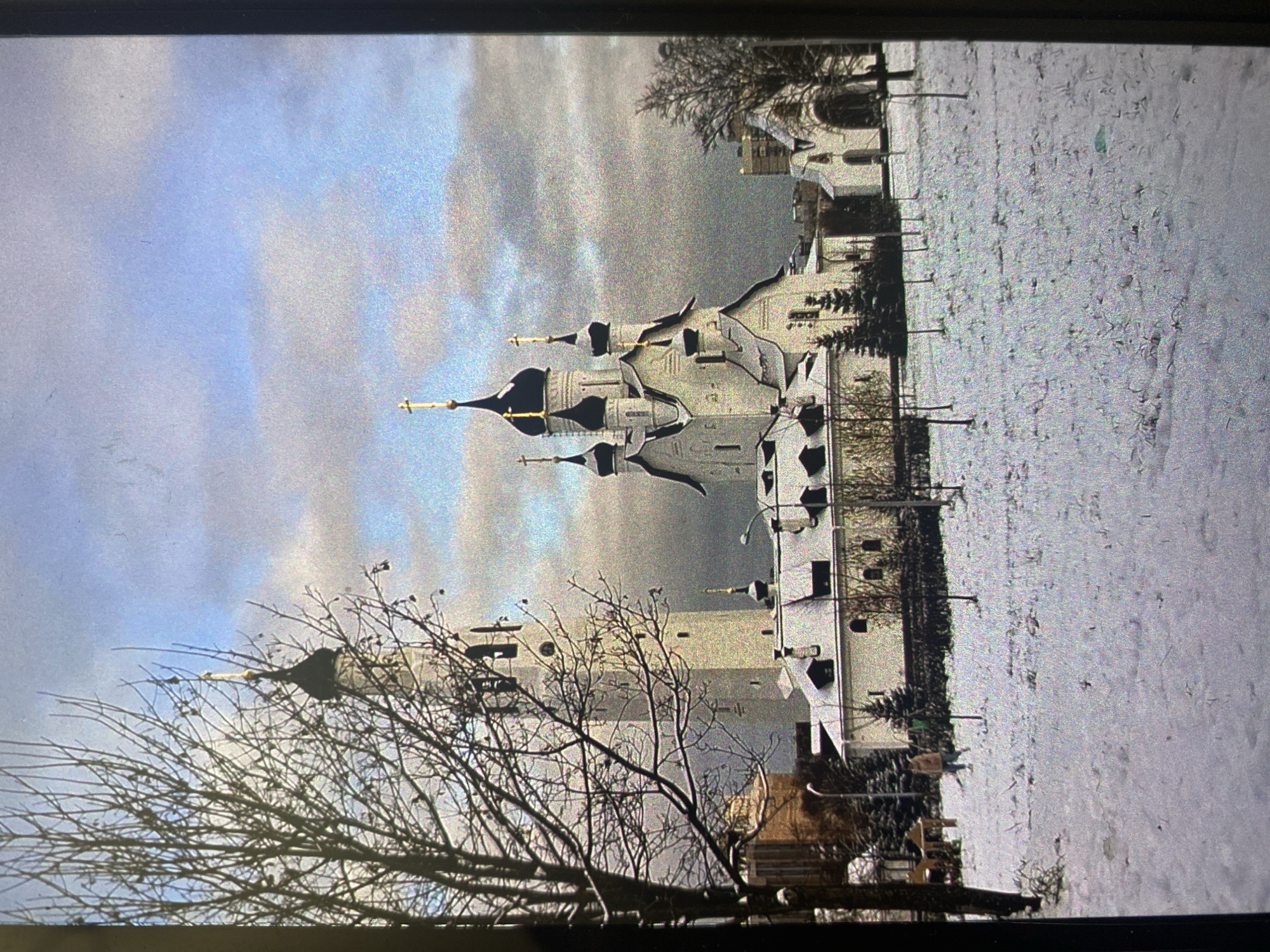 This media file, appearing to be a photograph of a screen, depicts a winter scene primarily featuring a grand Eastern Orthodox church. The image, though rotated clockwise, showcases the church with its distinctive multiple onion domes, each topped with a golden Orthodox cross, set against an overcast sky that blends pale blue and grey tones.
The scene is clearly set in winter, with the ground uniformly covered in a layer of snow. Bare-branched trees, devoid of leaves, frame parts of the church and extend into the foreground, reinforcing the cold season. Several other buildings, likely part of the same religious complex, are visible in the background and to the sides of the main church. A faint human figure can be observed walking across the snowy grounds in one of the cropped sections, providing a sense of scale to the expansive landscape.
The overall atmosphere conveys a sense of peaceful winter stillness and historical solemnity. The architectural style of the church, with its iconic domes, is characteristic of Russian Orthodox churches, aligning with the given location context of Saint Petersburg, Russia. There is no visible text present in the image.