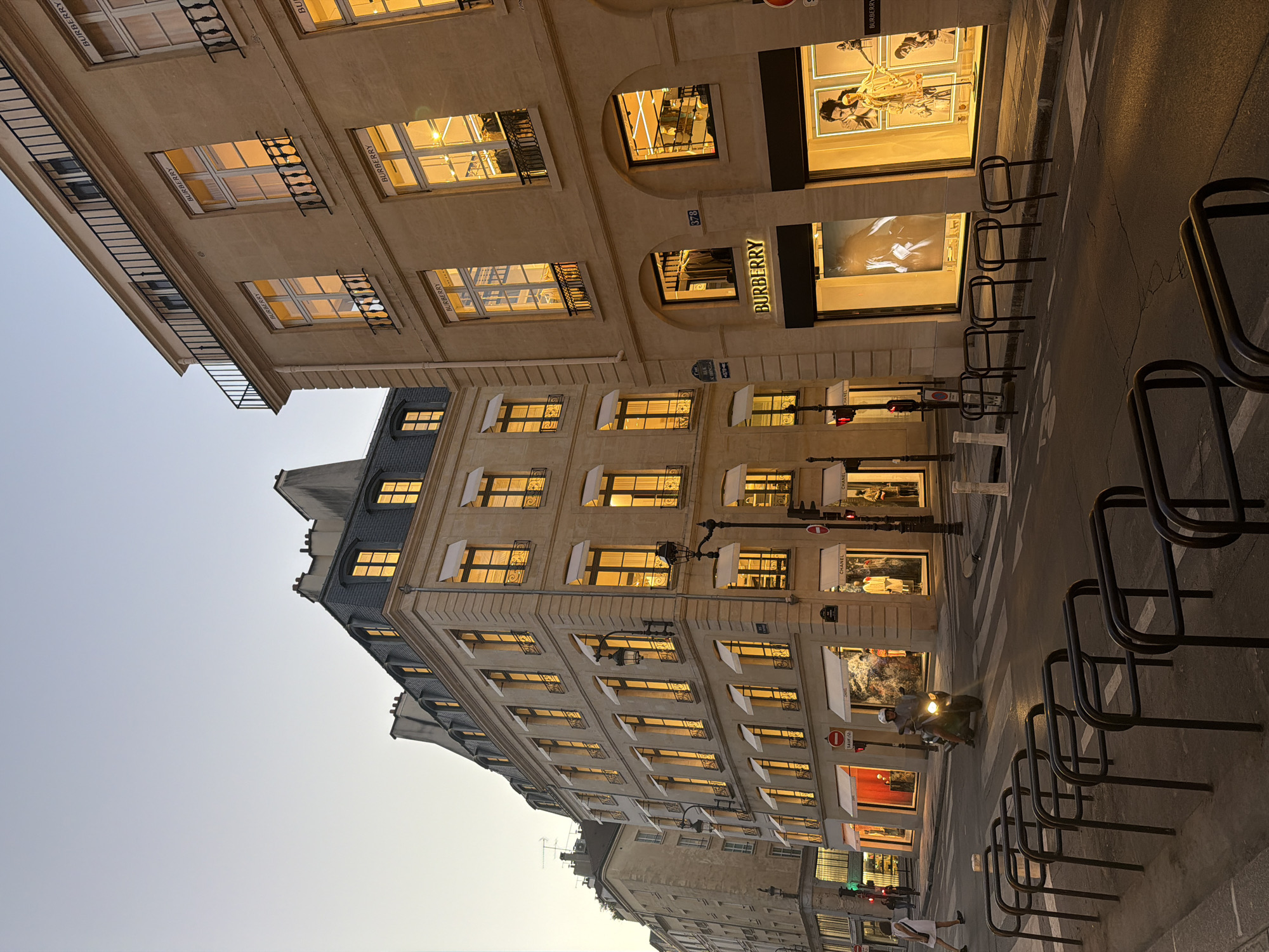 The image depicts a street scene in Paris, France. It is a sideways view showing a street with sidewalks on either side. A zebra crossing is visible, leading to the Burberry store. The building on the left is the Burberry store and the building across the street is the Chanel store.
There are several buildings in the image. They appear to be old buildings with similar architectural styles. They have many windows that are lit up. There are black metal racks along the sidewalk. A scooter is driving by in the road, and a woman is walking on the sidewalk. The time of day appears to be either early morning or late evening as the sky is light.