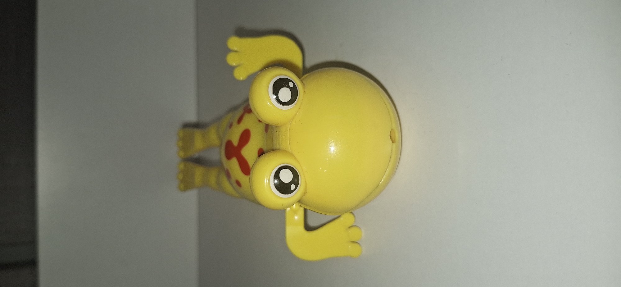 A yellow plastic toy frog is lying on its back on a white surface. The frog has large, round, cartoonish eyes with black pupils and white highlights. It has red spots on its back and limbs. The frog's legs are extended upwards, and its front limbs are bent outwards, with one raised as if waving. A small hole is visible on the underside of the frog's head. The background is a plain, light gray wall. There is no indication of the location in Bila Tserkva, Ukraine.