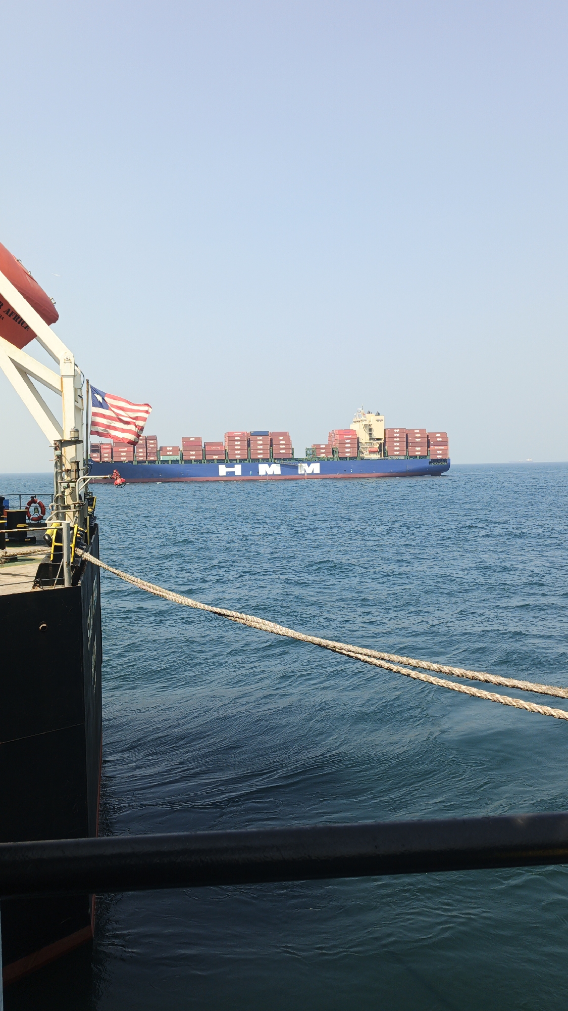 The image depicts a view from a ship at sea, likely in Singapore's waters, with a large container ship, the "HMM," prominently featured in the background. The container ship is loaded with red shipping containers stacked high on its deck. A Liberian flag is flown from the foreground vessel. Two thick ropes are secured, extending from the foreground ship towards the water, suggesting the ships are either docked or in the process of transferring cargo or personnel. The sea is calm with gentle ripples, and the sky is clear blue, indicating fair weather. The foreground shows a portion of a dark-colored ship, including a lifebuoy, and a black railing. No individuals are clearly visible in a manner that would allow for identification. The overall scene conveys a sense of maritime activity typical of a busy port.