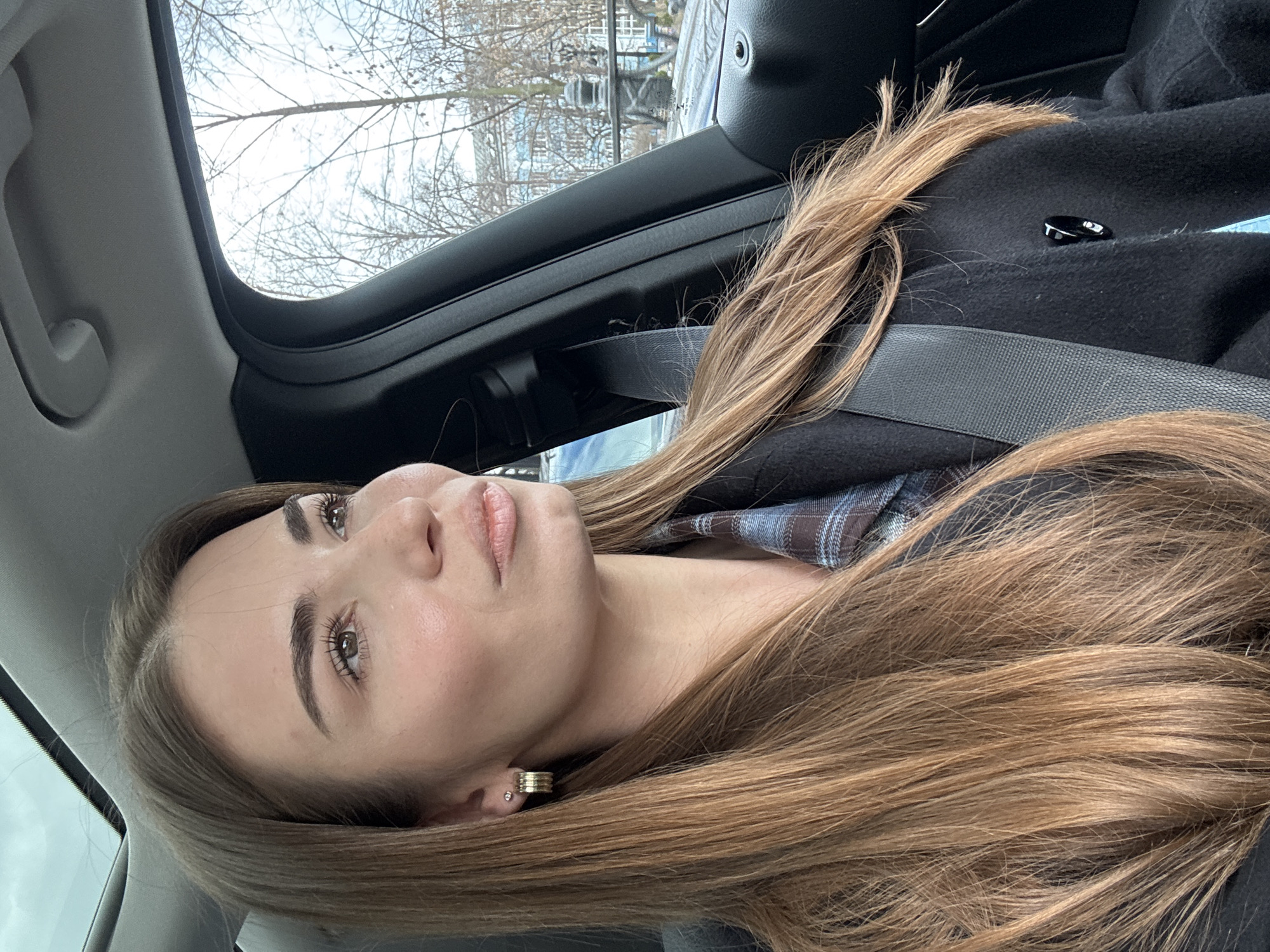 A young woman with long, light brown hair is seated in a car, wearing a dark coat and a plaid shirt, with the seatbelt fastened. She is looking upwards. The car interior is visible, including the window frame and the roof. Outside the window, bare trees and a building can be seen. There is no information to indicate if this image was taken in Moscow, Russia.