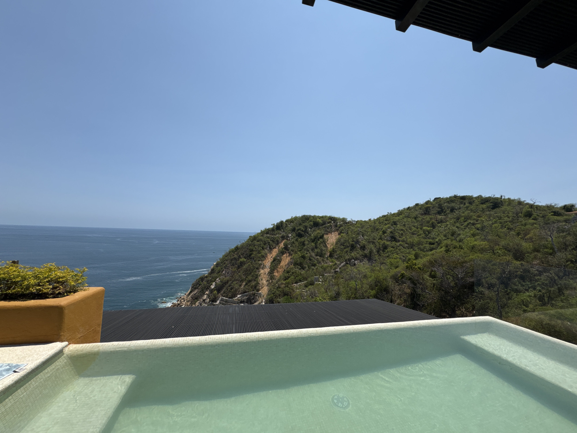 The image captures a view from a property in Puerto Marqués, Mexico. In the foreground is a light green tiled pool filled with water. Behind the pool is a black, corrugated roof. Beyond the roof, there's a lush green hill sloping down to the coast. The Pacific Ocean stretches to the left, meeting the horizon under a clear blue sky. A yellow-orange planter with green vegetation sits to the left of the roof. A section of a dark brown wooden overhang is visible at the top of the image.