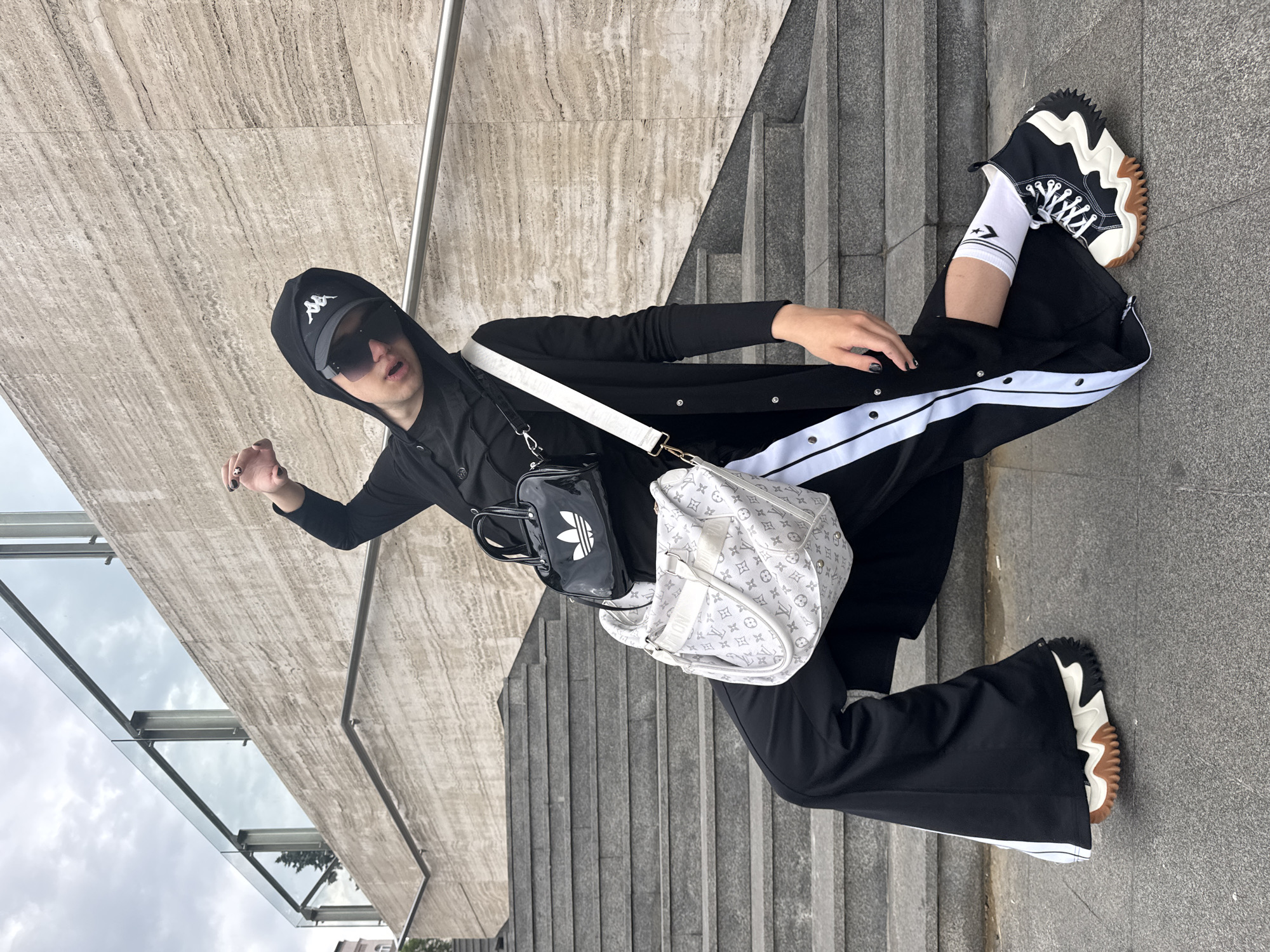 In Belgrade, Serbia, a person in a black hoodie, sunglasses, and a black baseball cap with a white logo stands on a concrete staircase with a textured stone wall behind them. They are wearing a black long coat with white stripes down the sides of the pants, white socks with a logo, and black high-top sneakers with white laces and a brown sole. The person is also carrying two bags: a small black patent bag with a white Adidas logo and a larger white patterned shoulder bag. They are striking a pose with one arm raised. The sky is overcast.