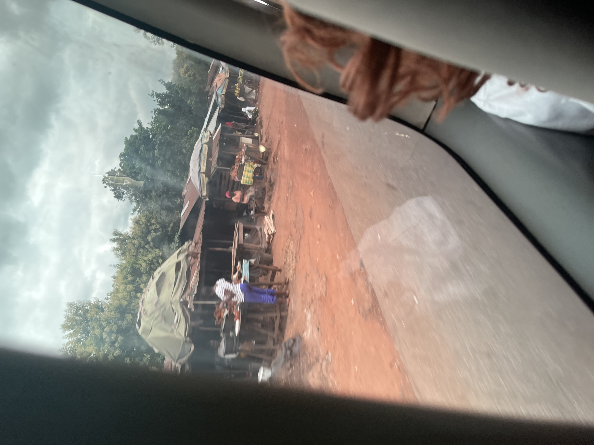 In Gidan Waya, Nigeria, this image captures a roadside market scene observed from inside a vehicle. The market features several stalls constructed with corrugated metal roofs and wooden supports, some covered with tarps. Items for sale, likely food or produce, are displayed on tables. Several individuals are visible, some tending to the stalls and others appearing to be customers. The ground is reddish-brown, suggesting dirt or laterite. In the foreground, the interior of the vehicle is visible, with part of a person's hair in the lower right corner. The sky is overcast.