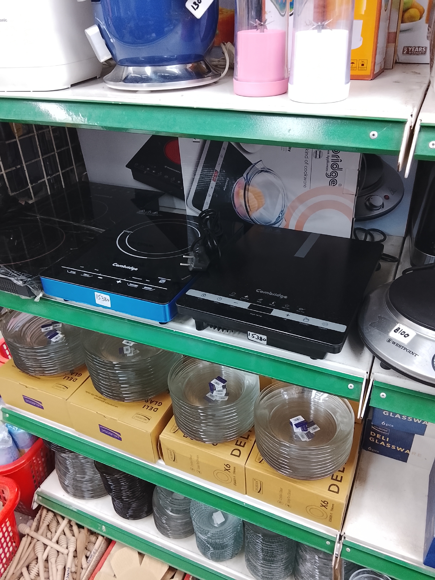 This is a cluttered shelf inside a store, likely in Gulgasht Colony, Pakistan, based on the image's origin. On the top shelf are kitchen appliances, including a white mixer, a blue kettle with a price tag of "130," and a pink and white blender. The next shelf holds two "Cambridge" brand induction cooktops, one with a blue frame and the other black, along with a "Westpoint" brand appliance. Both cooktops have price tags "15380" attached to them. Underneath, on the third shelf, are stacks of "DELI" brand glassware in boxes. The bottom shelf contains various utensils, including wooden honey dippers in a red basket. The shelves are green and supported by metal racks. The scene is typical of a small retail establishment selling household goods.