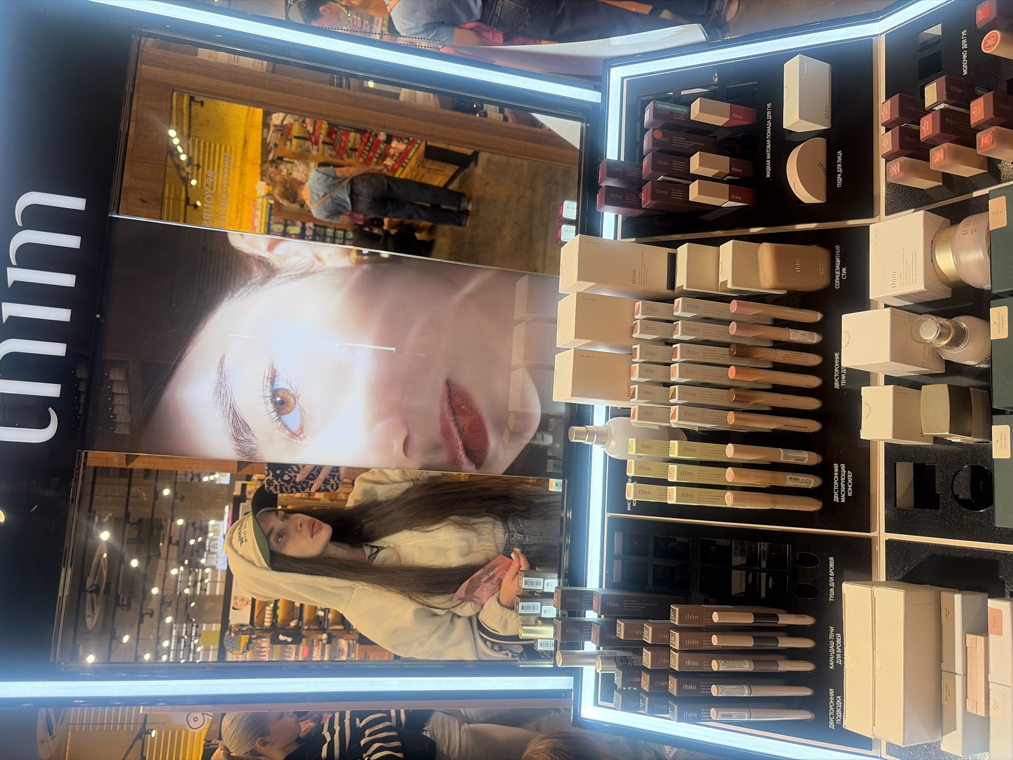 The image shows a brightly lit display of cosmetic products, predominantly from the brand "thim." The display is organized into multiple shelves and compartments, showcasing various items such as lipsticks, powders, concealers, and eyebrow products. The products are packaged in sleek, minimalist boxes and tubes, with "thim" branding visible on many of them.
In the reflection of the display, a young woman with long brown hair is visible. She is wearing a light-colored hoodie and a baseball cap, and is holding a phone with a leopard print case, possibly taking a selfie. Her gaze is directed slightly off-camera, and she appears to be in a retail environment, likely a beauty store or a section of a department store. The reflection also shows shelves stocked with more products in the background, and hints of wooden architectural elements.
The overall setting appears to be indoors, with artificial lighting casting a bright glow on the products. The presence of Russian text on some of the product packaging ("ЖИДКАЯ МАТОВАЯ ПОМАДА ДЛЯ ГУБ," "ПУДРА ДЛЯ ЛИЦА," "ТУШЬ ДЛЯ БРОВЕЙ," etc.) suggests the location is in Russia. The image captures a moment of browsing or shopping for cosmetics, with the woman interacting with the display or being captured in its reflection.