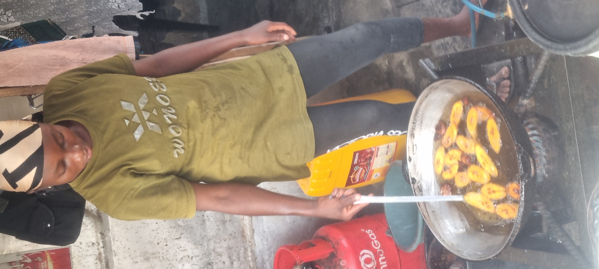 In Lagos, Nigeria, a person stands by a stove, frying plantains in a large metal pan filled with hot oil. The person is wearing a green t-shirt with a graphic design and a patterned headscarf. To the left of the pan is a red gas cylinder, and to the right is a large yellow container. The background shows a rough concrete wall and some hanging fabrics. The overall scene suggests a street food vending or home cooking environment.