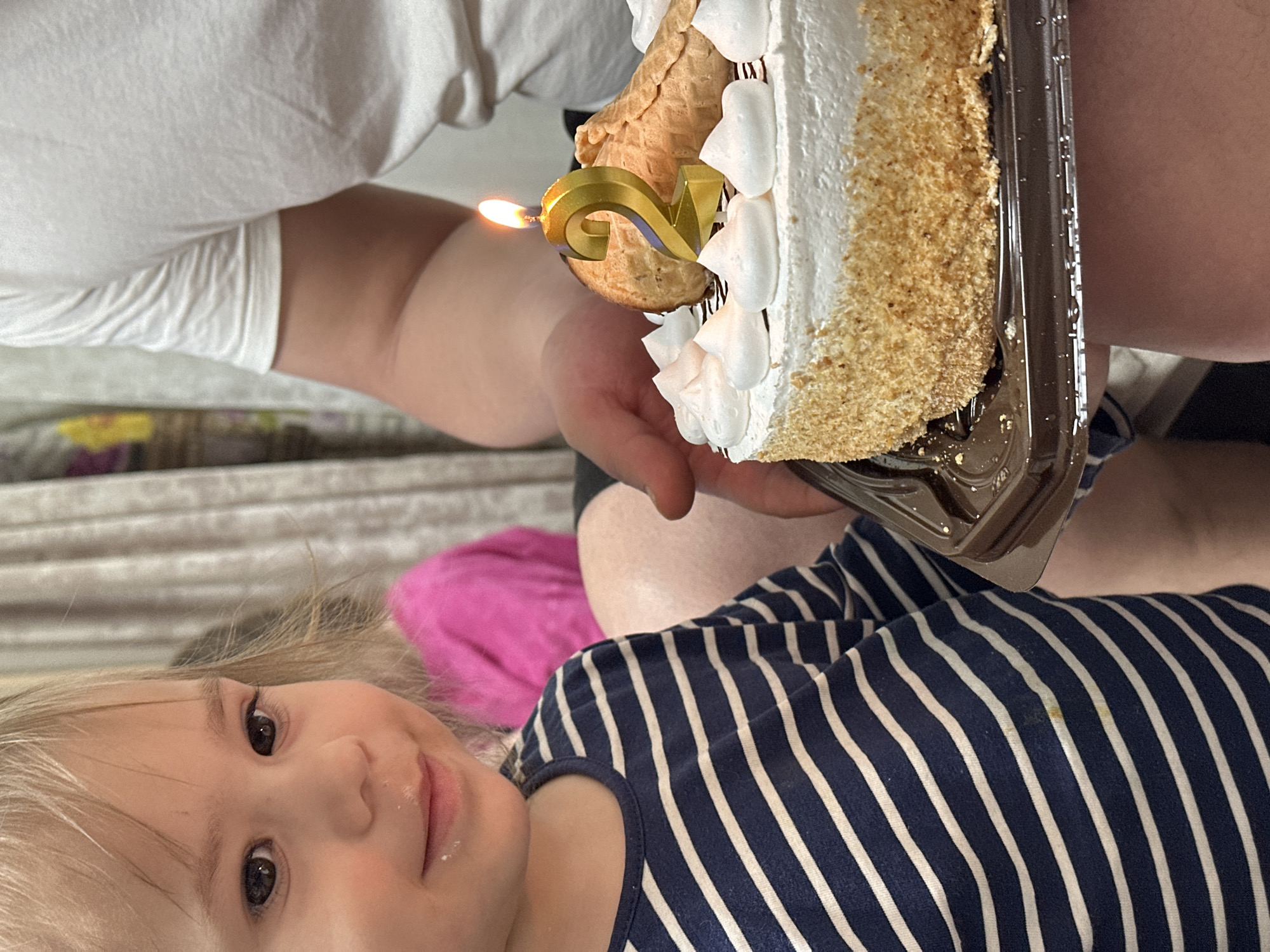 A young child with fair hair and a stripey blue and white shirt is positioned in the foreground. Their face is turned towards the viewer, and there is a small amount of white substance around their mouth. A cake with white frosting and a lit "2" candle sits above and behind the child. The cake is being held by a person with a white shirt. The background features a patterned fabric and a pink object, but these are not sharply defined. The location is Uva, Russia.