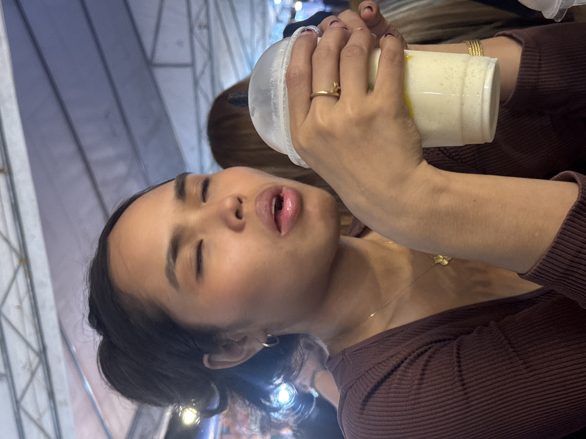A close-up, slightly tilted shot captures a woman holding a drink and about to take a sip. Her eyes are closed, and her mouth is open, suggesting she's in the process of drinking. The drink appears to be a creamy, light-colored beverage in a clear plastic cup with a clear lid. She is wearing a maroon, ribbed long-sleeved shirt and several pieces of jewelry, including a gold ring on her right hand and a thin gold necklace with a pendant. A gold bracelet is also visible on her left wrist. The background is blurred, but hints of a structure with crisscrossing supports and lights suggest an indoor or covered outdoor setting, possibly an event or market. The overall lighting suggests it could be evening. No visible text is present. The image was taken in Davao City, Philippines.