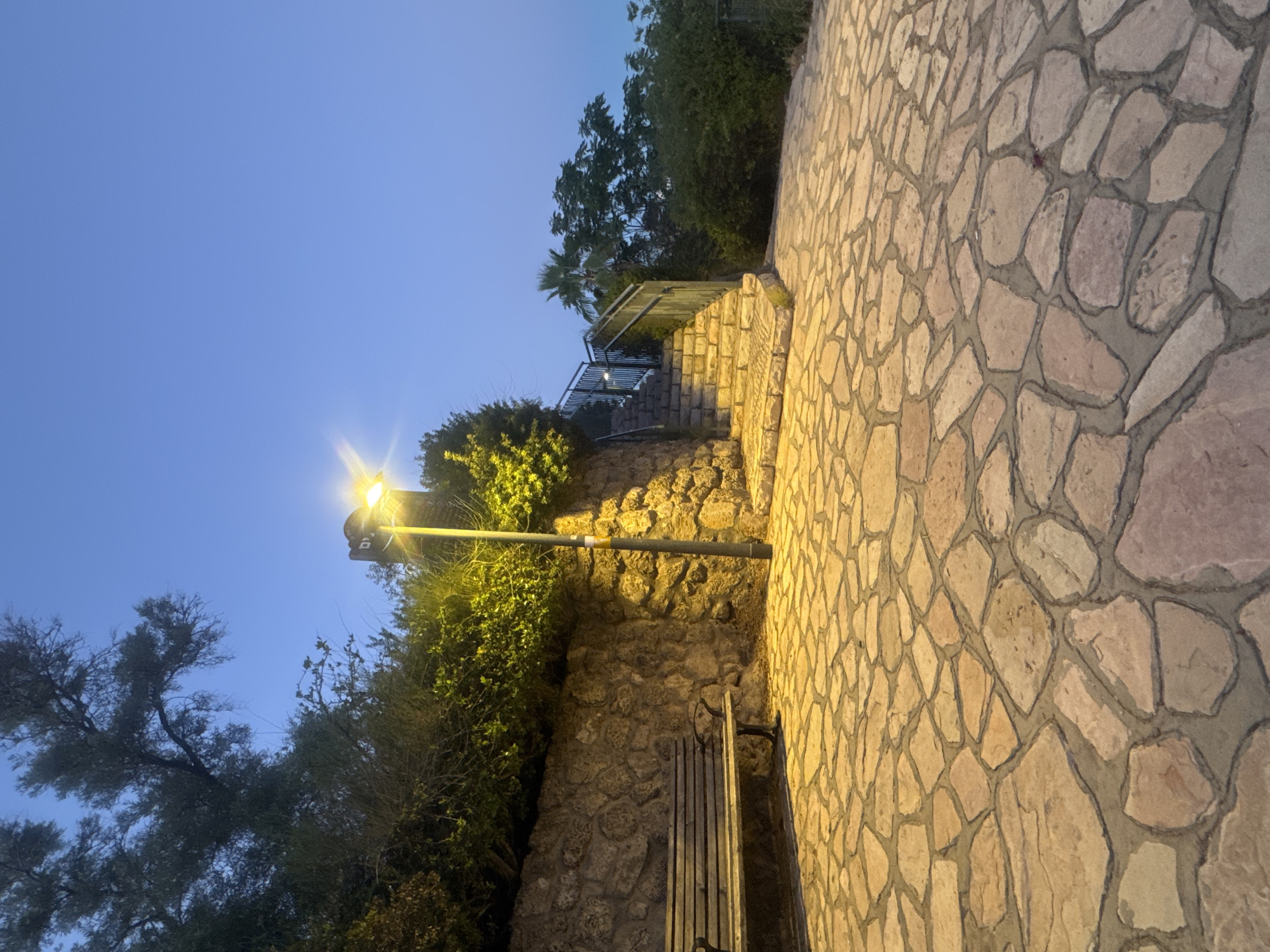 The image shows a park pathway and stairway in Ramat Gan, Israel, at dusk. The pathway is paved with irregular, flat stones. To the left, a wooden bench sits in front of a stone wall covered with green foliage. A lamppost with a bright light illuminates the scene. A stone stairway with metal railings leads up and to the right, bordered by greenery. The sky is a clear, deep blue, indicating early evening. The overall impression is of a peaceful, well-maintained outdoor public space.