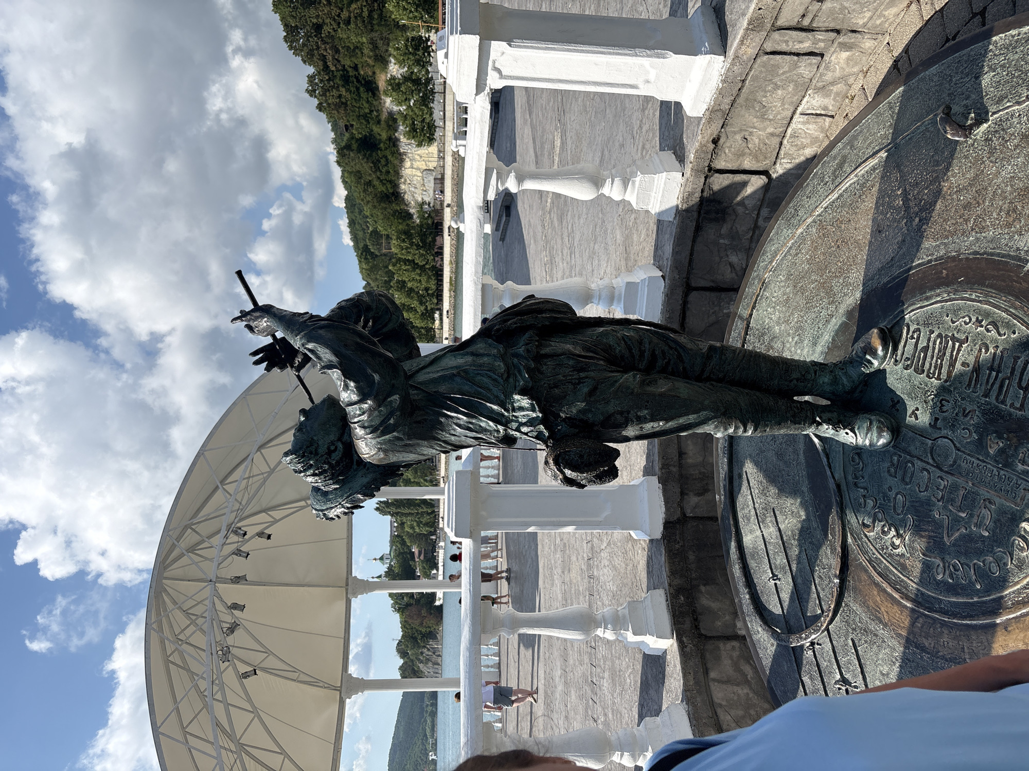 In Abrau-Dyurso, Russia, a bronze statue of a man playing a flute stands on a circular base in an outdoor area. The statue is dark green with hints of patina. The man is depicted wearing loose-fitting clothes and has a bag slung over his shoulder. He is holding a long, thin flute to his lips. The base of the statue features engraved text and circular patterns.
In the background, a white, open-air gazebo with a domed roof and supporting columns is visible. Beyond the gazebo, a body of water, likely a lake or bay, stretches out. On the far side of the water, lush green hills rise, dotted with trees. A building with a green dome is also visible on the hillside. A white railing with decorative posts lines the promenade in front of the gazebo, and several people are walking along it. A person wearing a light blue top is partially visible on the left side of the frame, in the foreground. The sky is a bright blue with scattered white clouds. The scene suggests a pleasant day in a scenic, public outdoor space.