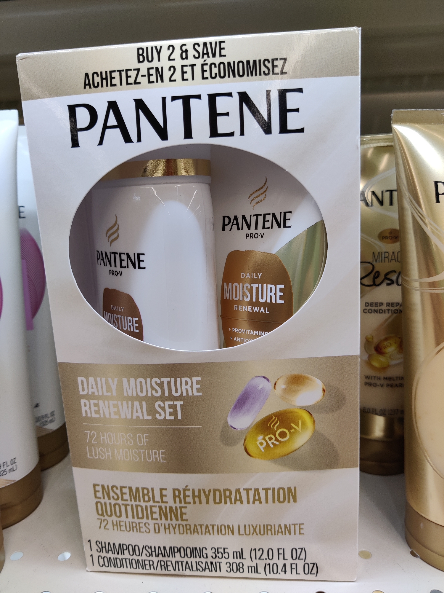 The image shows a retail display shelf in a store. The main subject is a Pantene Daily Moisture Renewal Set gift set, which is a boxed product containing two hair care items. The box is white and gold and features large "PANTENE" branding. Inside the box, two bottles are visible, one appearing to be a shampoo and the other a conditioner. The set is advertised with "BUY 2 & SAVE" and "ACHETEZ-EN 2 ET ÉCONOMISEZ" promotions. There are other Pantene products on the shelf to the right and left of the gift set, including what appears to be a Miracle Rescue deep repair conditioner. The products are arranged on a white shelf. No people are visible in the image. The lighting suggests an indoor retail environment. The location of Mechanicsburg, United States of America, is not explicitly indicated in the visual information presented.
