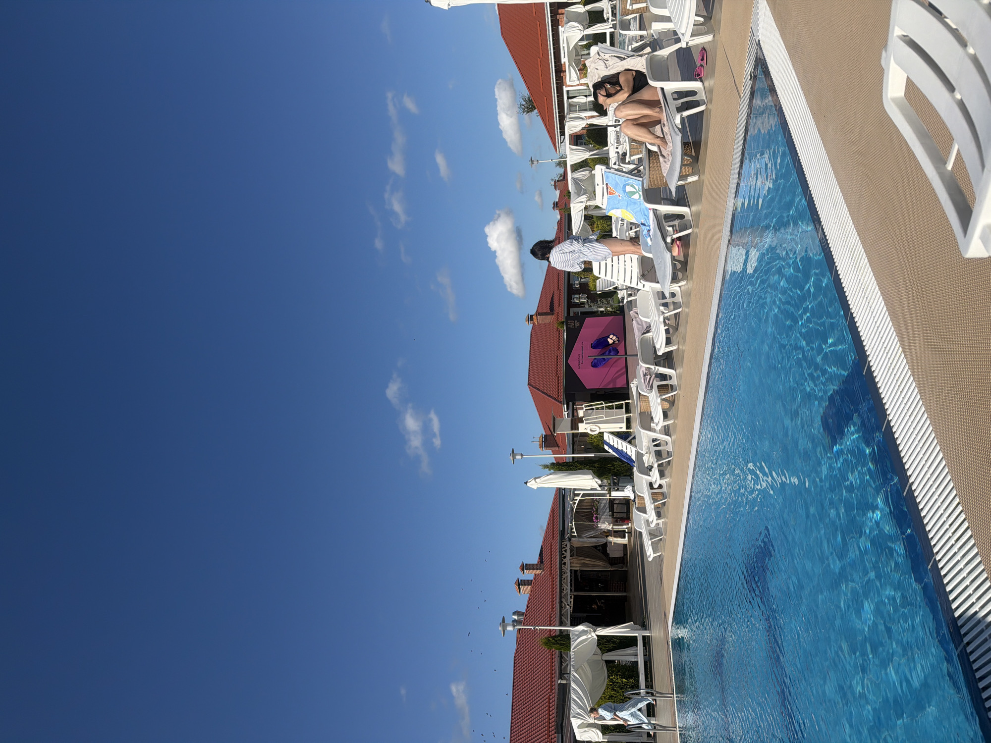 A bright, sunny day at an outdoor swimming pool facility is depicted. The pool itself is a vibrant blue, with sunlight reflecting off its surface. Surrounding the pool is a beige deck. In the background, there are several buildings with red tiled roofs, some featuring white canopy structures. A line of white lounge chairs is visible along the pool's edge.
There are two women in the scene. One woman, wearing a blue and white striped shirt and dark bottoms, is standing near a lounge chair. The second woman, wearing a black swimsuit, is reclining on a lounge chair, holding what appears to be a phone or camera to her face. There are also several white lounge chairs, some unoccupied and others with towels or bags on them. A few potted plants and trees are scattered throughout the scene, adding greenery to the environment. The sky is a clear, bright blue with a few scattered white clouds. A flock of birds can be seen flying in the distance against the sky. The image appears to be taken in Laskino, Russia, suggested by context not visible in the image itself.