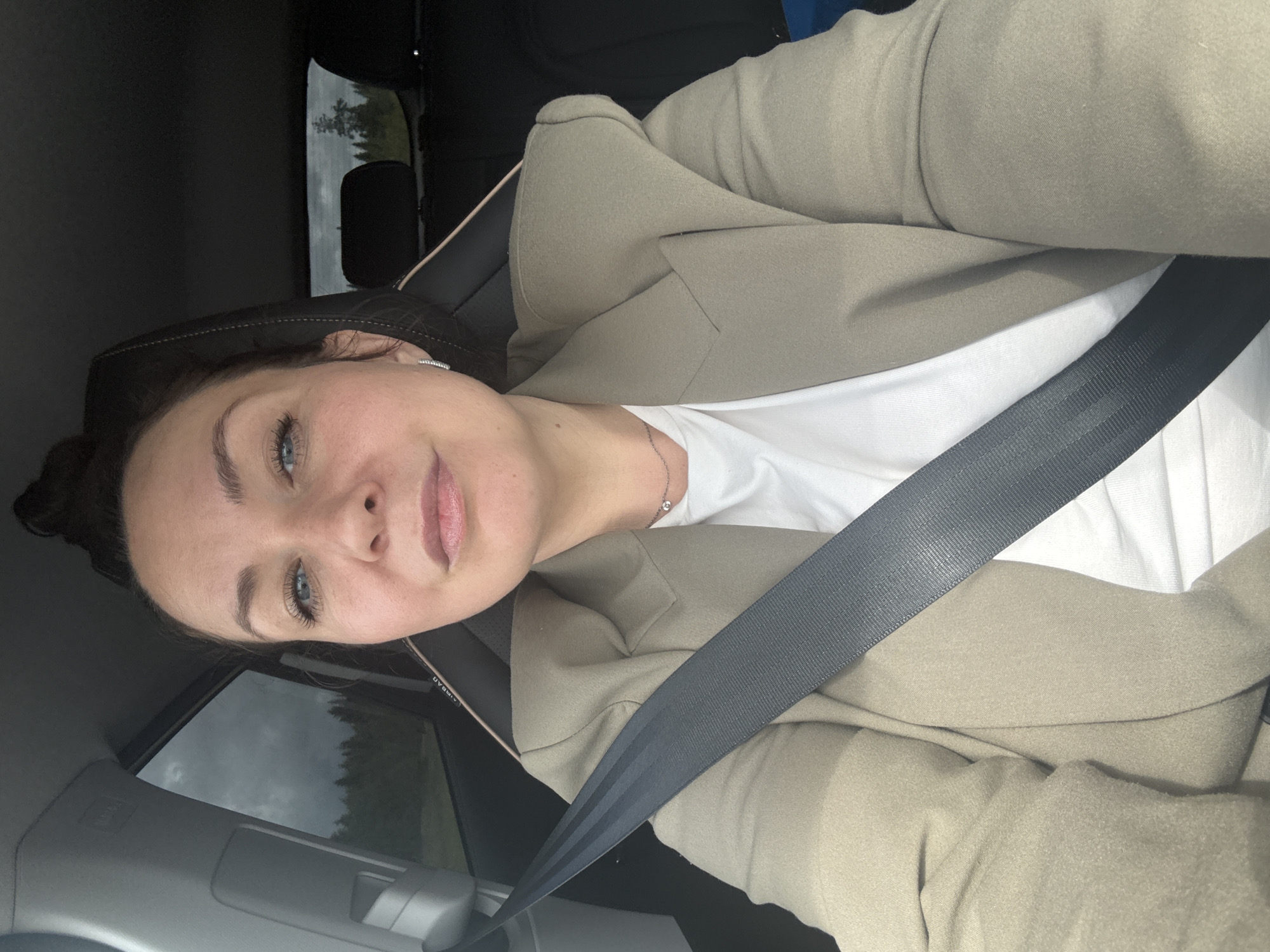 A woman is captured in a selfie taken from inside a car, likely in the passenger seat, in Kukushtan, Russia. She has dark hair, blue eyes, and a light complexion, exhibiting a calm and slightly smiling expression. She is wearing a white t-shirt, a light tan or beige blazer, and a delicate silver necklace with a small pendant. A black seatbelt is fastened across her chest.
Through the car window, an overcast sky is visible above a stretch of dark green trees, consistent with a rural or wooded setting. The interior of the car is dark, with visible seat upholstery and a headrest. Partially visible and inverted text that appears to read "AIRBAG" can be seen on the car's interior, possibly on a headrest or pillar. The time of day is daytime, characterized by cloudy or overcast weather.