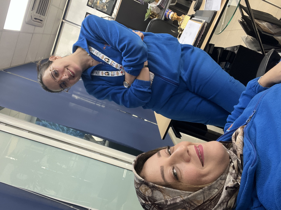 This image features two women in an indoor office setting. The woman in the foreground, positioned to the left, is wearing a patterned headscarf and a royal blue zippered fleece jacket, looking towards the camera with a smile. Her right arm, adorned with a silver-colored bracelet, is visible. Standing behind her, slightly to the right, is a second woman wearing glasses, a royal blue zippered fleece jacket, and matching blue pants. She is also smiling, with her arms crossed, and has a lanyard with multiple small badges around her neck, and a watch on her left wrist. A light-colored desk with a black frame is visible between and behind the women, holding a black desk pad, papers, a golden bust of Nefertiti, and a decorative plant. The background consists of a light blue wall partition, a large glass partition with white framing and vertical blinds, and a white ceiling equipped with an air conditioning vent. The precise location within the city of Meknes, Morocco, cannot be determined from the visual information provided.