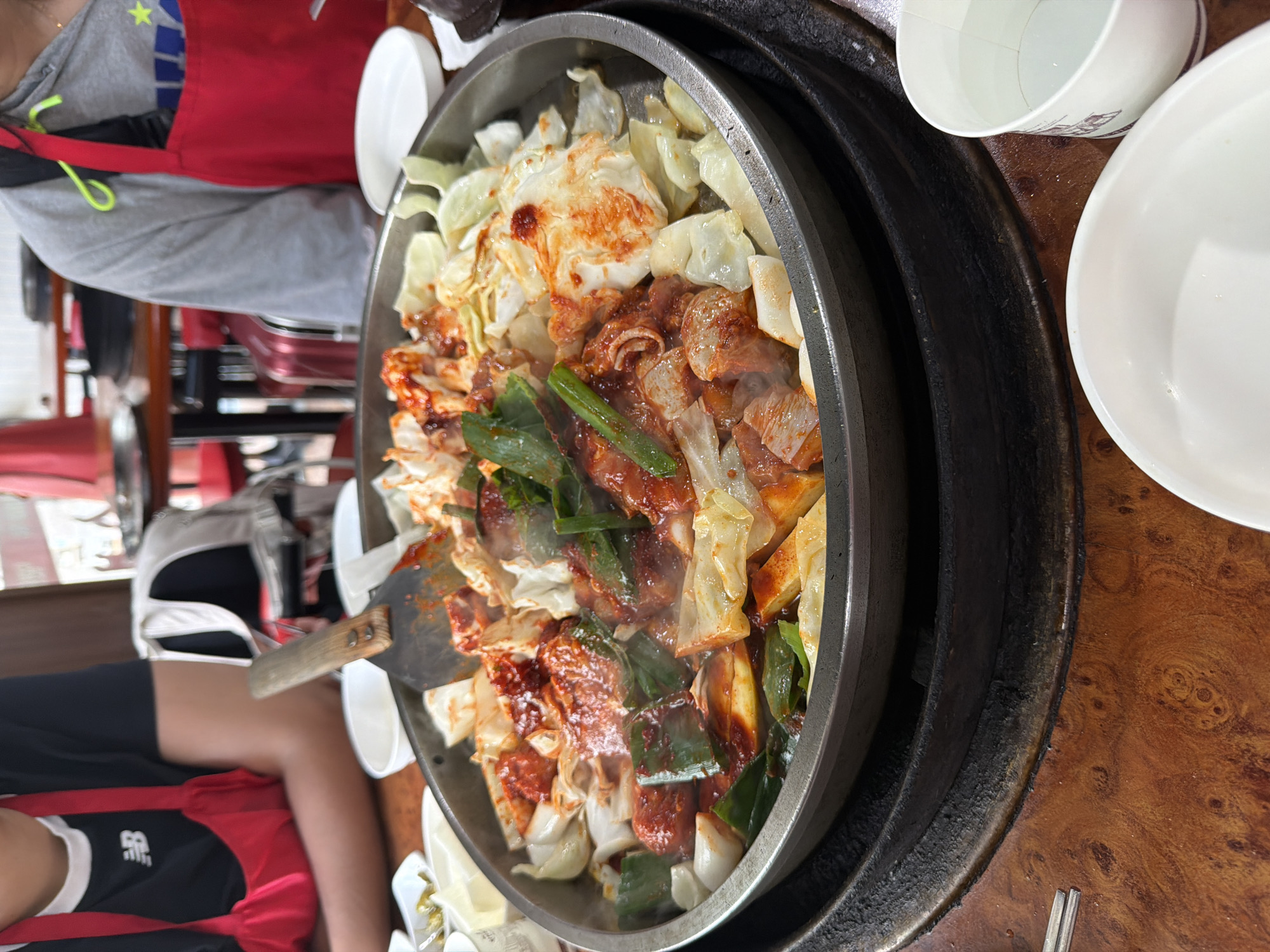 In a casual dining establishment in Chuncheon-si, South Korea, a close-up shot captures a vibrant dish of dakgalbi, a popular spicy stir-fried chicken dish, being prepared or served. The food is presented in a wide, shallow metal pan, likely over a heat source, with steam rising from the ingredients. The dakgalbi contains chunks of chicken, stir-fried with generous amounts of red chili paste sauce, shredded cabbage, green onions, and possibly tteok (rice cakes). A metal spatula rests in the pan, suggesting recent activity in mixing or serving the dish.
Two individuals are partially visible in the background. On the left, a person wearing a black t-shirt with a white 'NB' logo and a red apron is seated. Their arms and torso are visible, and they appear to be looking towards the food. On the right, another person, wearing a grey hoodie and a red apron, is also seated, with their upper body and arm visible. They are positioned further away from the camera, and their focus is not entirely clear.
The table is made of dark, wood-grained material, possibly a laminate or a treated wood surface, with distinct circular patterns visible. Several white ceramic bowls and a white paper cup filled with a clear liquid are placed around the main dish. The overall setting suggests a lively and informal dining experience, common in restaurants specializing in this type of cuisine in Chuncheon, a city renowned for its dakgalbi.