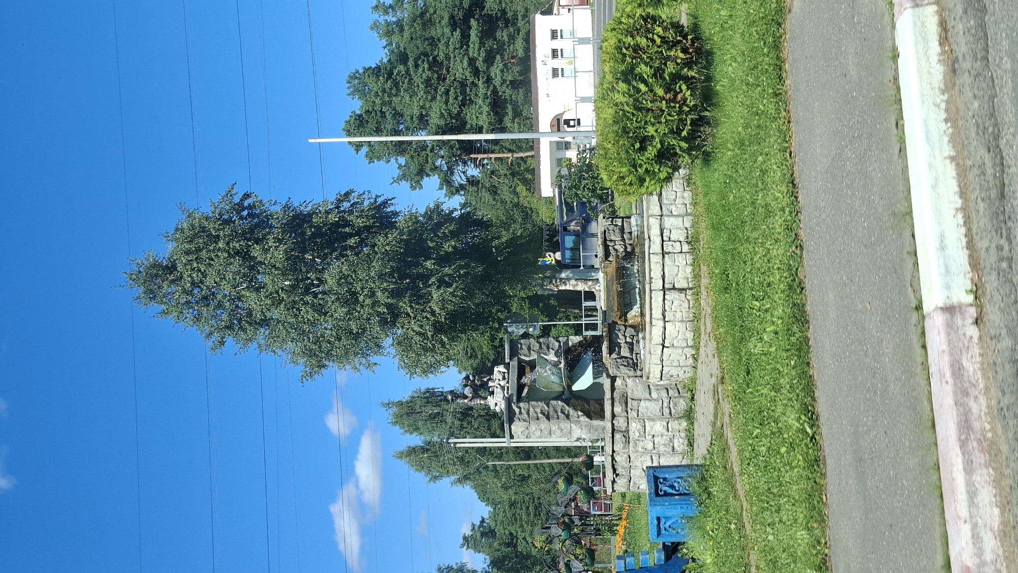 A medium shot captures a public fountain in the city of Pastavy, Belarus, on a bright, sunny day. The fountain, constructed from stacked stone blocks, features multiple tiers with flowing water. At the apex of the fountain, a sculpture of a boy and a dog is visible. The fountain is surrounded by green grass and a trimmed hedge. In the background, a building with white walls and several windows stands, and a dark-colored van is parked nearby. Large trees, including a prominent birch tree, frame the scene, and power lines stretch across the clear blue sky. To the left of the fountain, there are two blue benches or planters. The foreground shows a paved road with a colored curb.