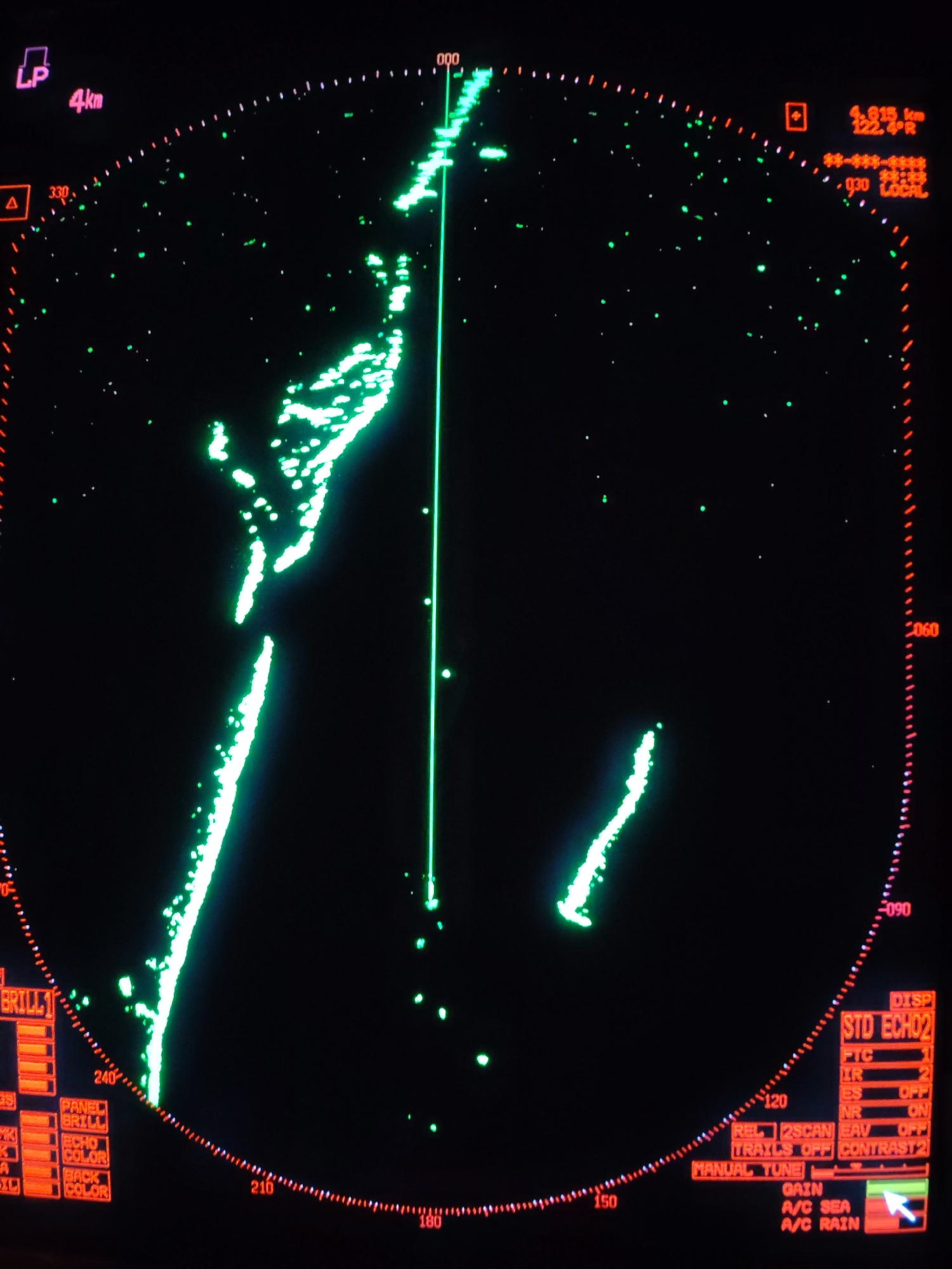 This image shows a radar screen, likely from a ship. The screen displays green echoes on a black background, indicating the presence of objects or terrain. A vertical green line marks the ship's heading. A cluster of echoes on the left side of the screen might represent land or a large vessel. A smaller cluster on the lower right suggests another object. Red text and markings around the screen's perimeter provide information like distance (4.615 km), bearing, and radar settings. The controls are labeled with terms like "GAIN", "BRILL", and "CONTRAST". The top left displays "LP 4km" and the top right displays distance and bearing information. There's no information to suggest this is related to Kostyaevka, Russia.