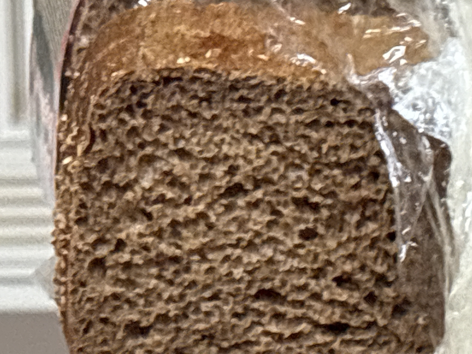 The image shows a close-up of a slice of dark bread, likely rye or pumpernickel, with a visible crumb structure. The bread is slightly moist and appears to be enclosed in clear plastic packaging. The background is blurred and out of focus, suggesting the bread is on a surface with some vertical elements behind it. There is no indication of the city of Georgiyevsk, Russia, or any people, actions, or interactions in this image.