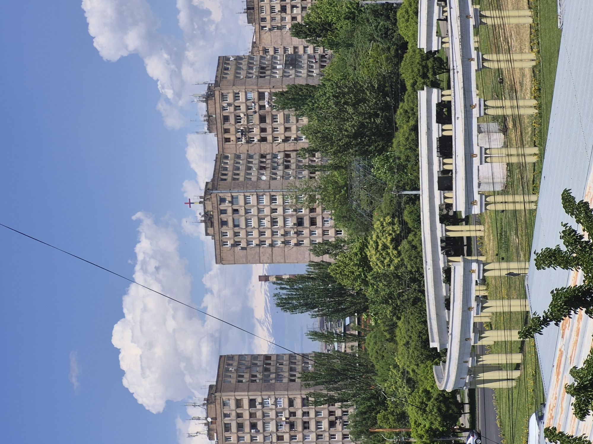 This image captures a bright, sunny daytime scene of an urban landscape, likely in Yerevan, Armenia. The photo is rotated 90 degrees clockwise, but when viewed upright, it features a clear blue sky with large, white cumulus clouds across the top.
Several prominent, multi-story apartment buildings, characteristic of Soviet-era concrete architecture, dominate the mid-ground. These buildings are a light grey or brown hue, featuring numerous windows and some balconies. Many of them are adorned with various satellite dishes and antennas on their roofs and facades. A notable red cross symbol is visible near the top of one of these buildings, possibly indicating a medical facility. In the distance, between two of the apartment blocks, a tall, slender industrial chimney rises, with faint, hazy outlines of mountains visible beyond, potentially Mount Ararat.
Abundant green trees with dense foliage are scattered throughout the scene, interspersed between the buildings and forming green buffers, suggesting a tree-lined city.
In the foreground, on the right side of the upright image, a distinctive elevated structure runs horizontally. This light-colored structure, possibly an aqueduct, a walkway, or a monument, features a series of yellowish-brown, surfboard-like vertical elements underneath. Below this structure, numerous dark utility wires or cables crisscross the frame. A partial view of a corrugated metal roof, showing patches of rust, is visible in the bottom right corner, and a narrow paved road can be seen with a small, dark car on it.
The scene depicts a calm, static urban environment under bright, clear daylight, suggesting midday or early afternoon. No specific activity or visible text is present.