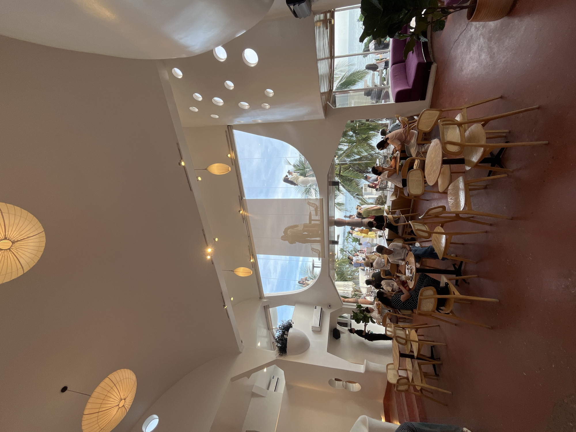 The image shows the interior of a brightly lit, modern cafe in Mũi Né, Vietnam, with a view of the ocean and palm trees through large windows. The cafe features a unique architectural design with curved white walls, circular cutouts, and large, open spaces. Several groups of people are seated at tables, engaged in conversation or dining. In the foreground, tables and chairs are arranged on a reddish-brown floor. In the midground, patrons are seated at various tables, some interacting with each other and others looking out at the view. In the background, the ocean and a clear sky are visible, with palm trees framing the scene. The overall ambiance suggests a relaxed and social atmosphere.