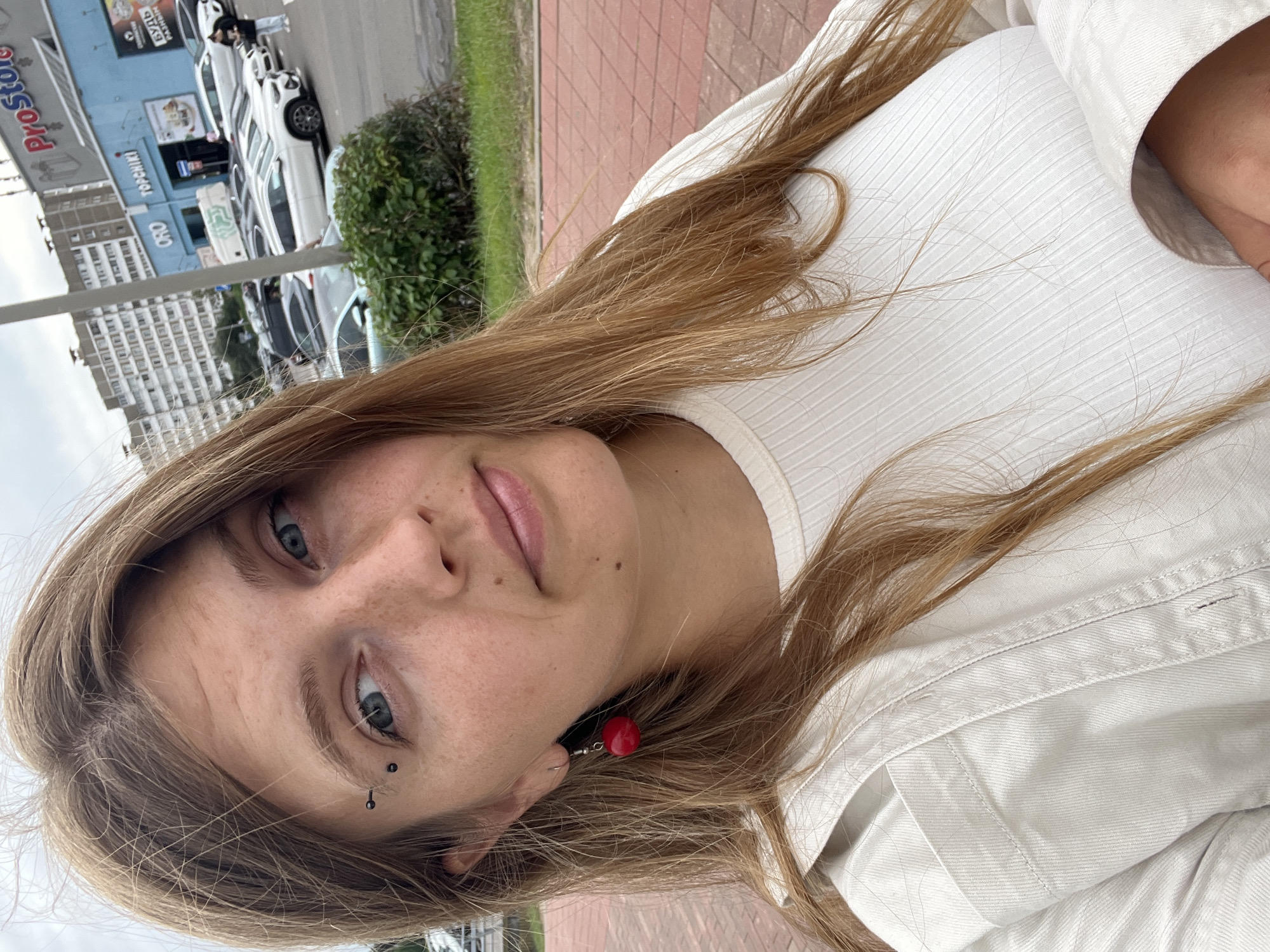 A medium shot photograph captures a young Caucasian woman with long, light brown hair and blue eyes. She is wearing a white ribbed top and a white jacket, with a red beaded earring in her left ear and an eyebrow piercing. The background suggests an urban environment, possibly a parking lot outside a shopping center. A building with blue and white signage, reading "ProStore" and other text in Cyrillic, is visible, along with parked cars. The ground in the foreground is paved with red bricks. The sky appears overcast.